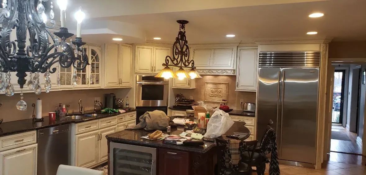 Cluttered kitchen with cream cabinets, stainless fridge, chandelier, and a large island under warm lighting