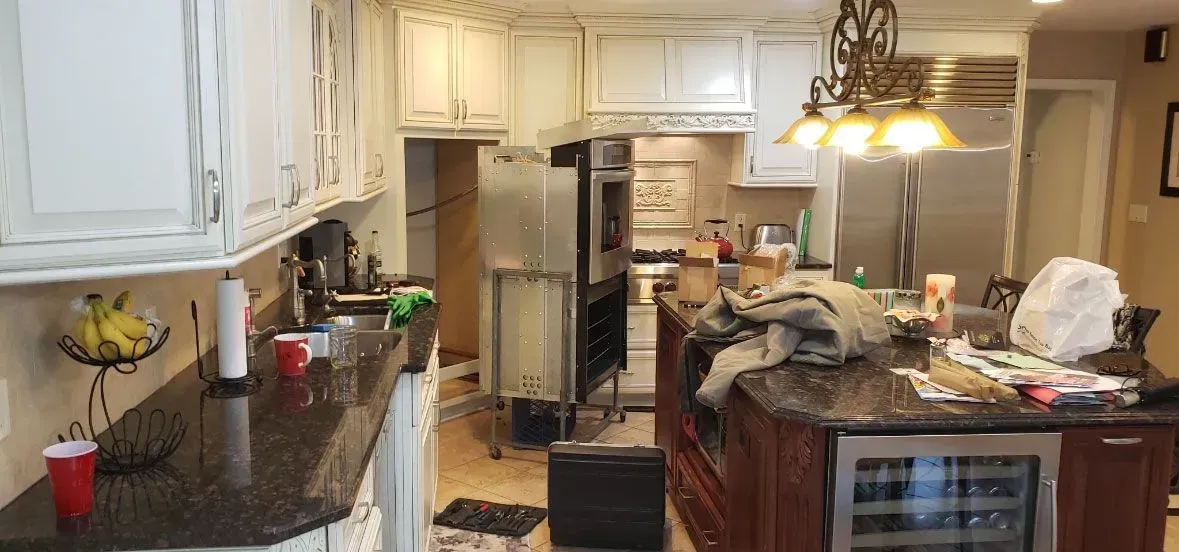 Cluttered kitchen with white cabinets, black countertops, and a central island covered in items.
