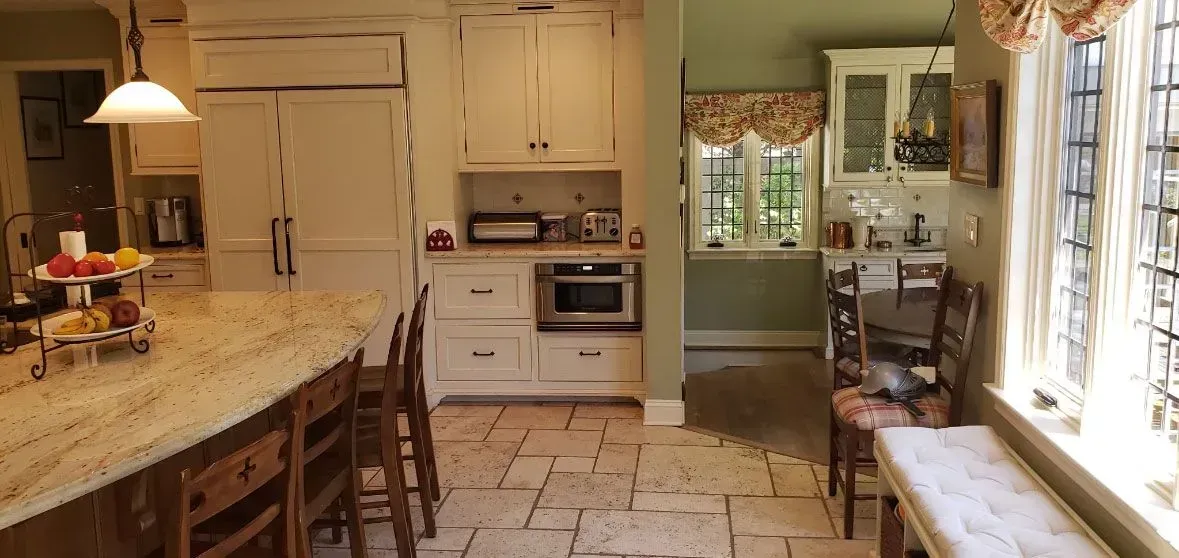 Bright kitchen with granite island, white cabinets, and large windows along a sunlit tiled floor