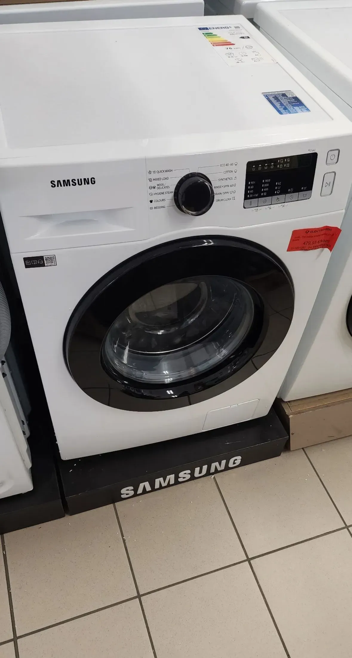 White front-loading washing machine in a store, with black door and control panel.