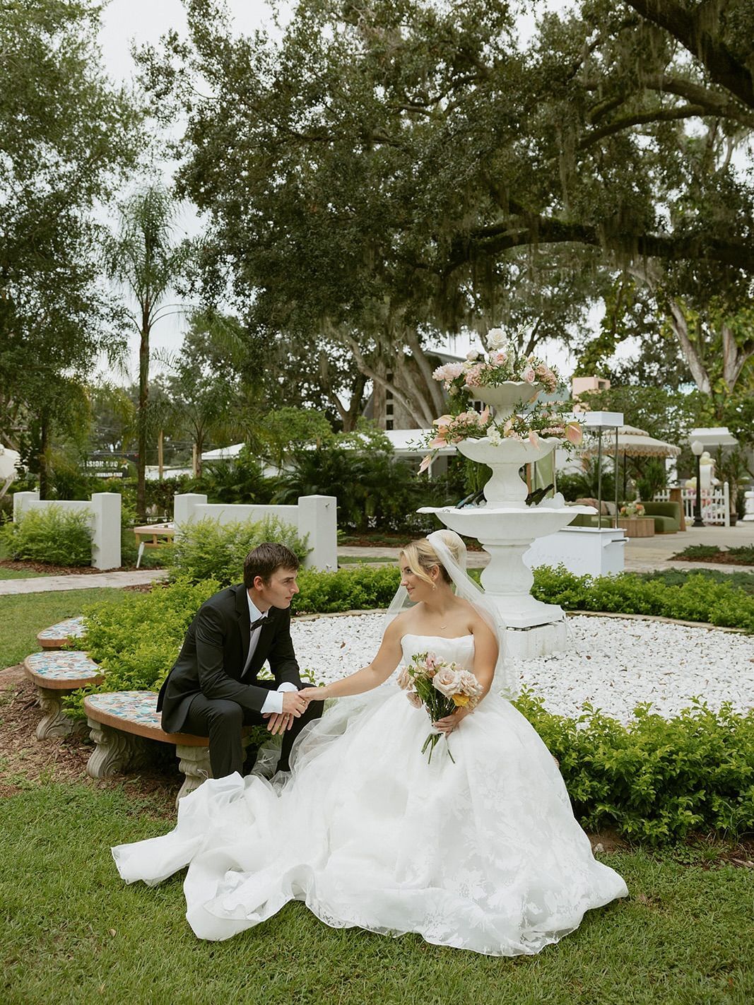 Bride and groom holding hands, sitting on a bench in a garden with a fountain in the background.