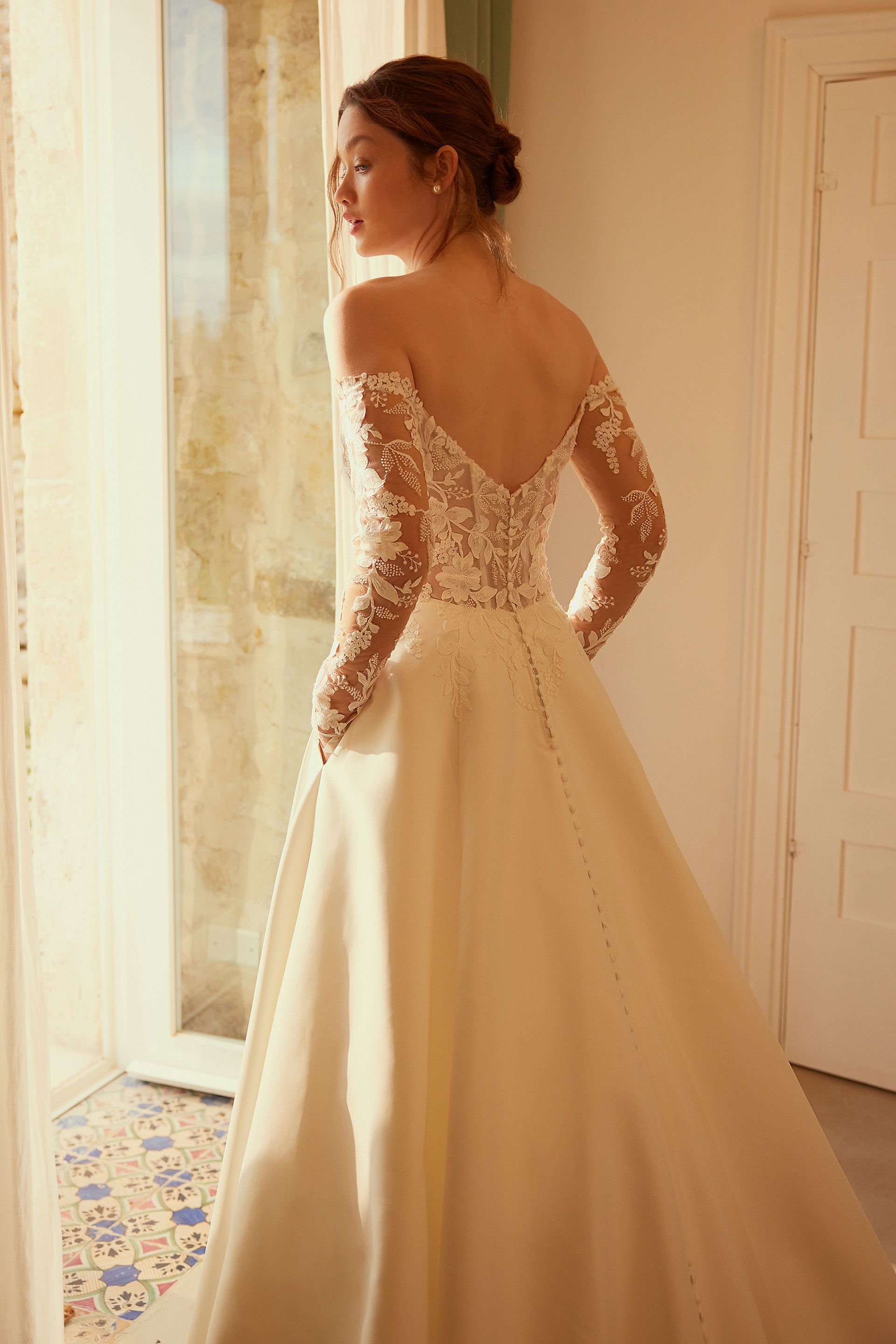 Woman in a wedding dress looks out a window. Dress is off-the-shoulder, white, with long sleeves and a full skirt.
