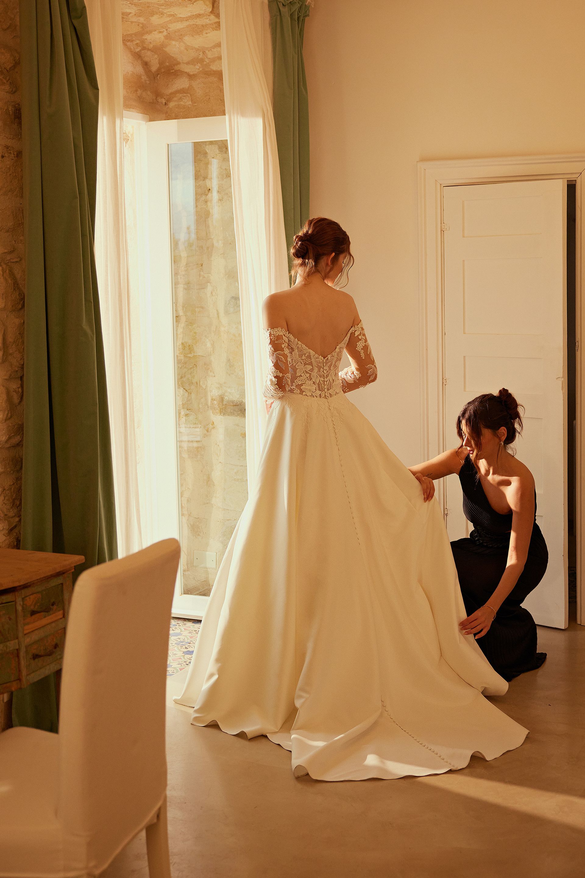 Bride in wedding dress, being helped by attendant. Sunlit room, dress is white with lace sleeves, attendant wears black.