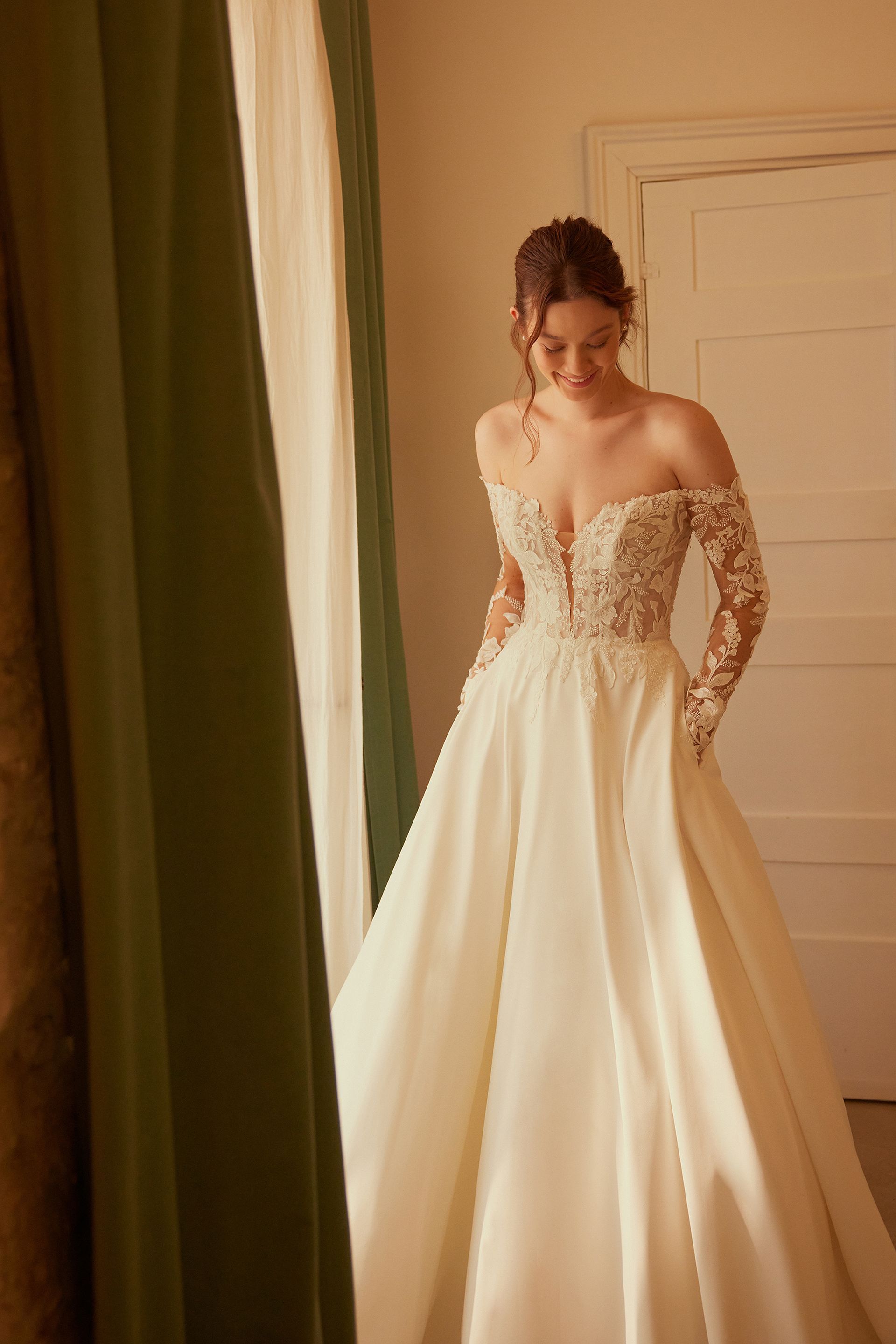 Woman in wedding dress, smiling, indoors. Off-shoulder bodice with floral embroidery, long sleeves, full skirt.