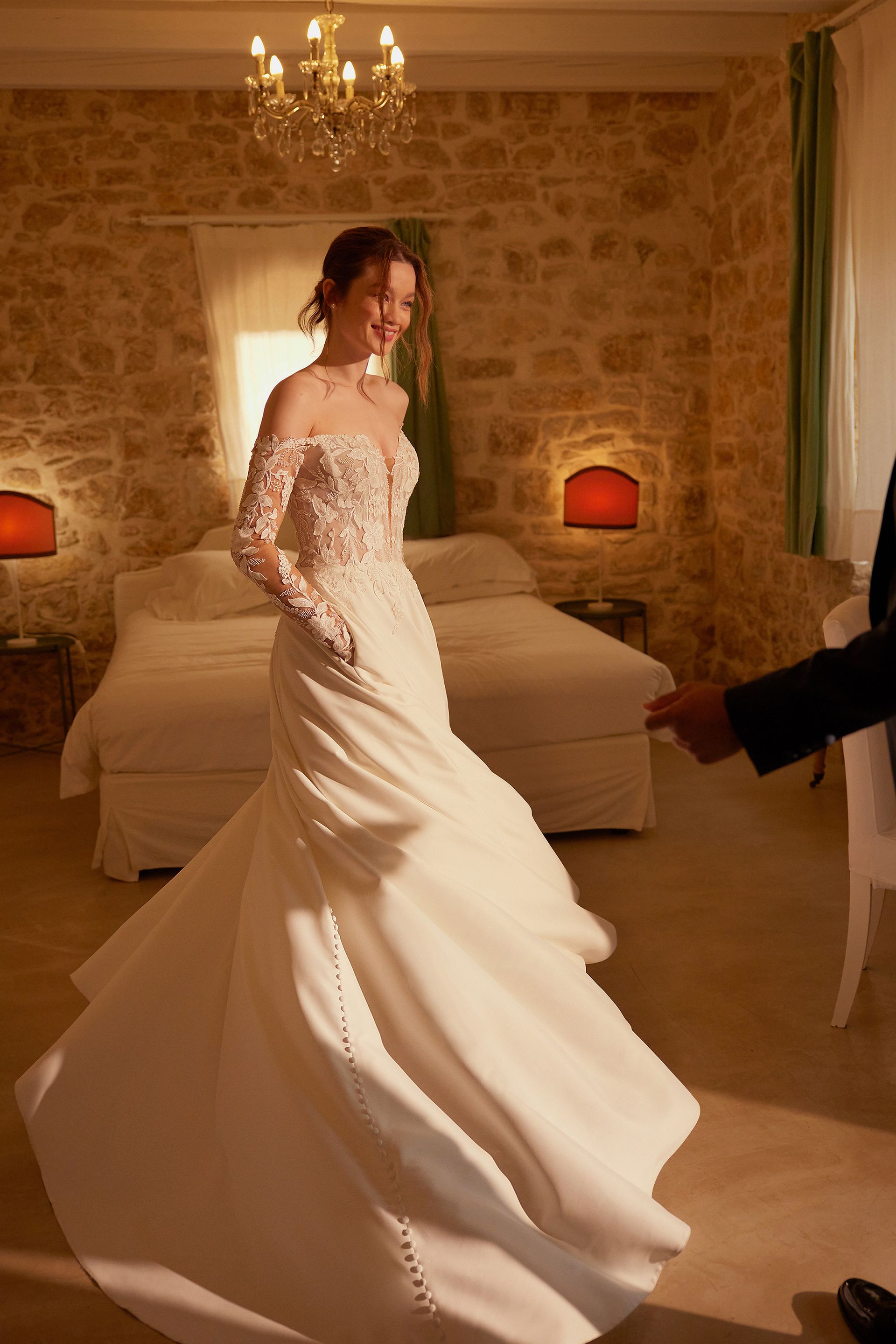 Bride in wedding dress, smiling in stone-walled room, near bed. Man's arm outstretched.