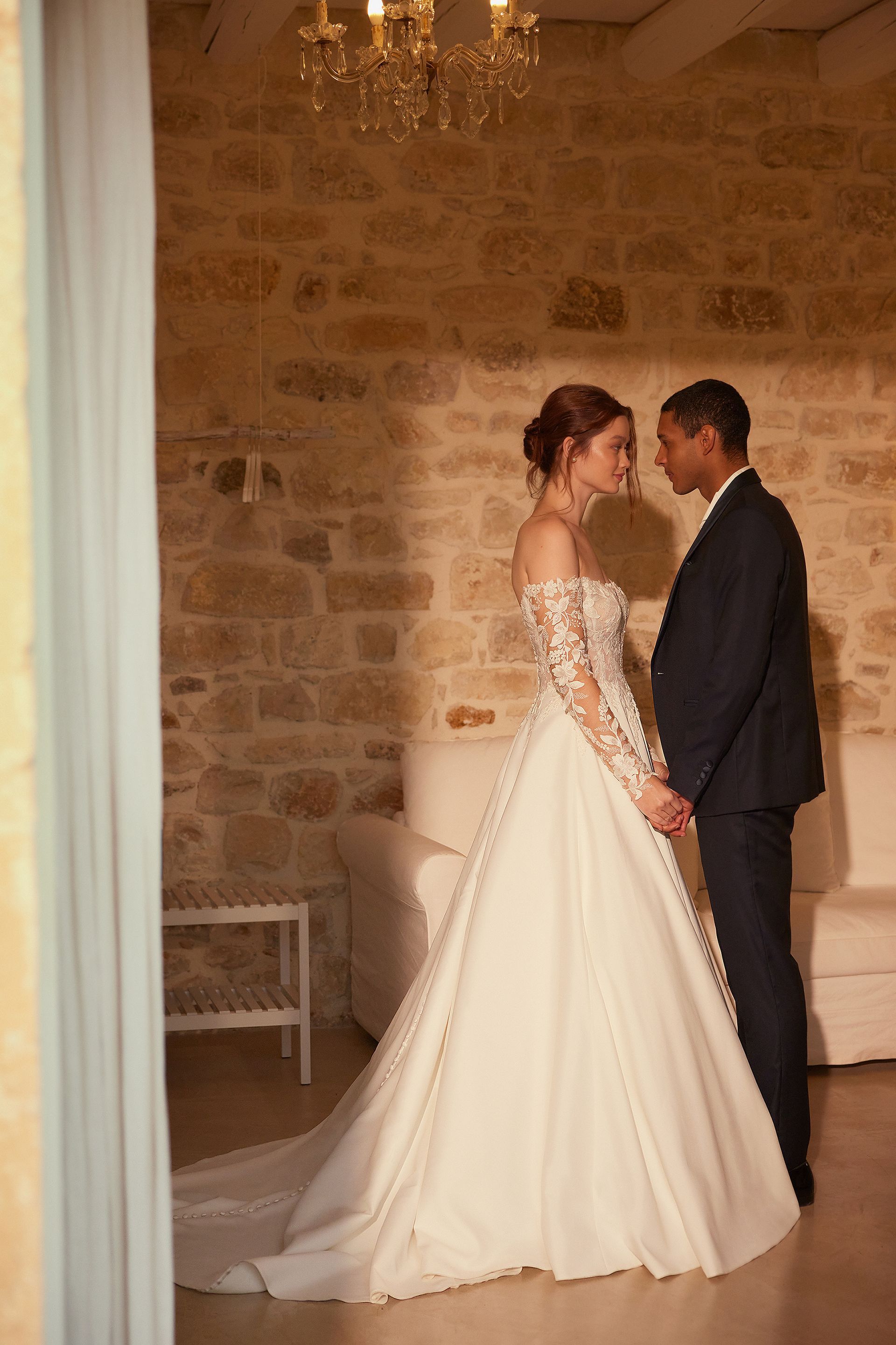 Couple holding hands, bride in white gown, groom in suit, standing in a stone-walled room.