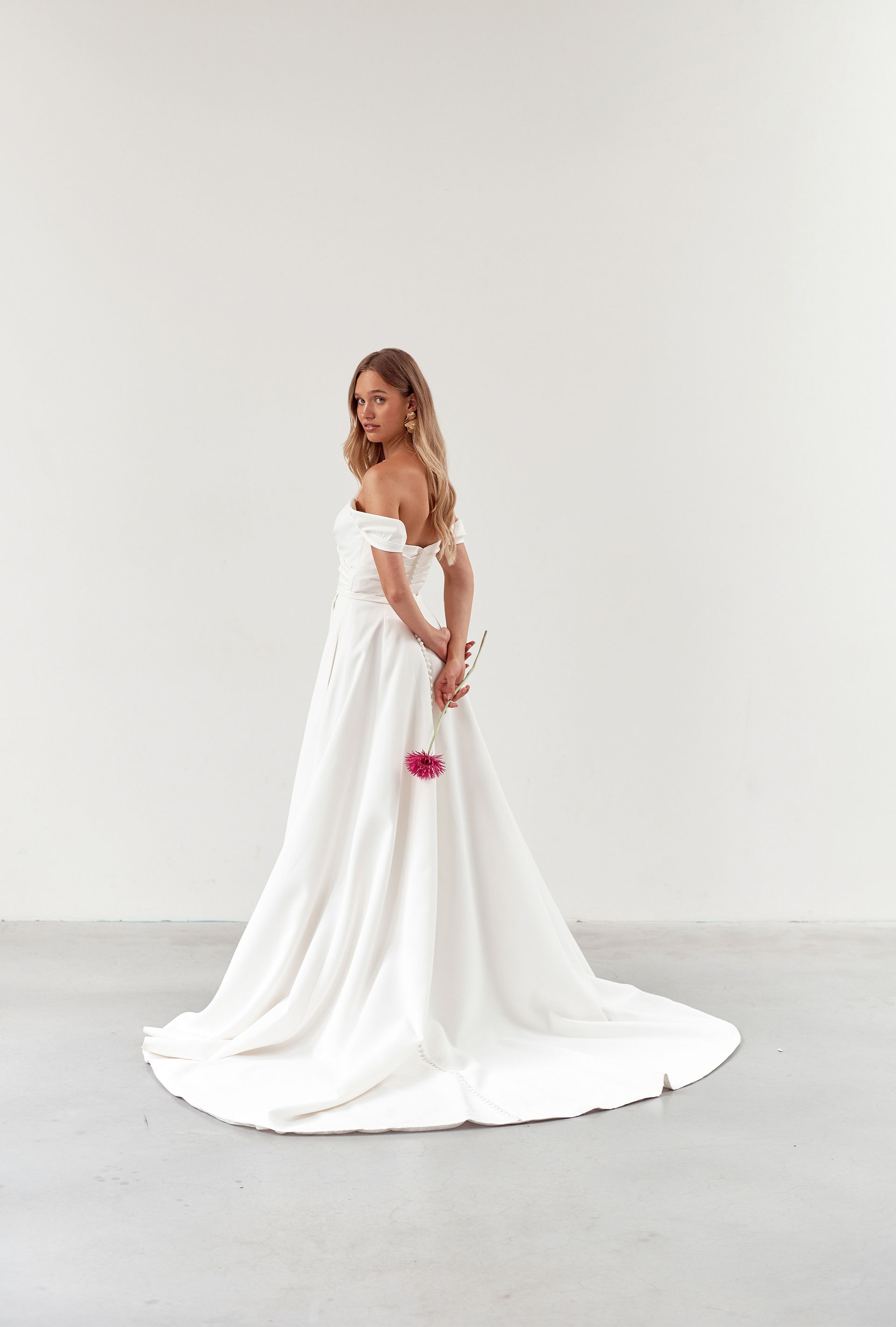 Woman in white off-shoulder wedding dress holding a pink flower, looking over her shoulder.