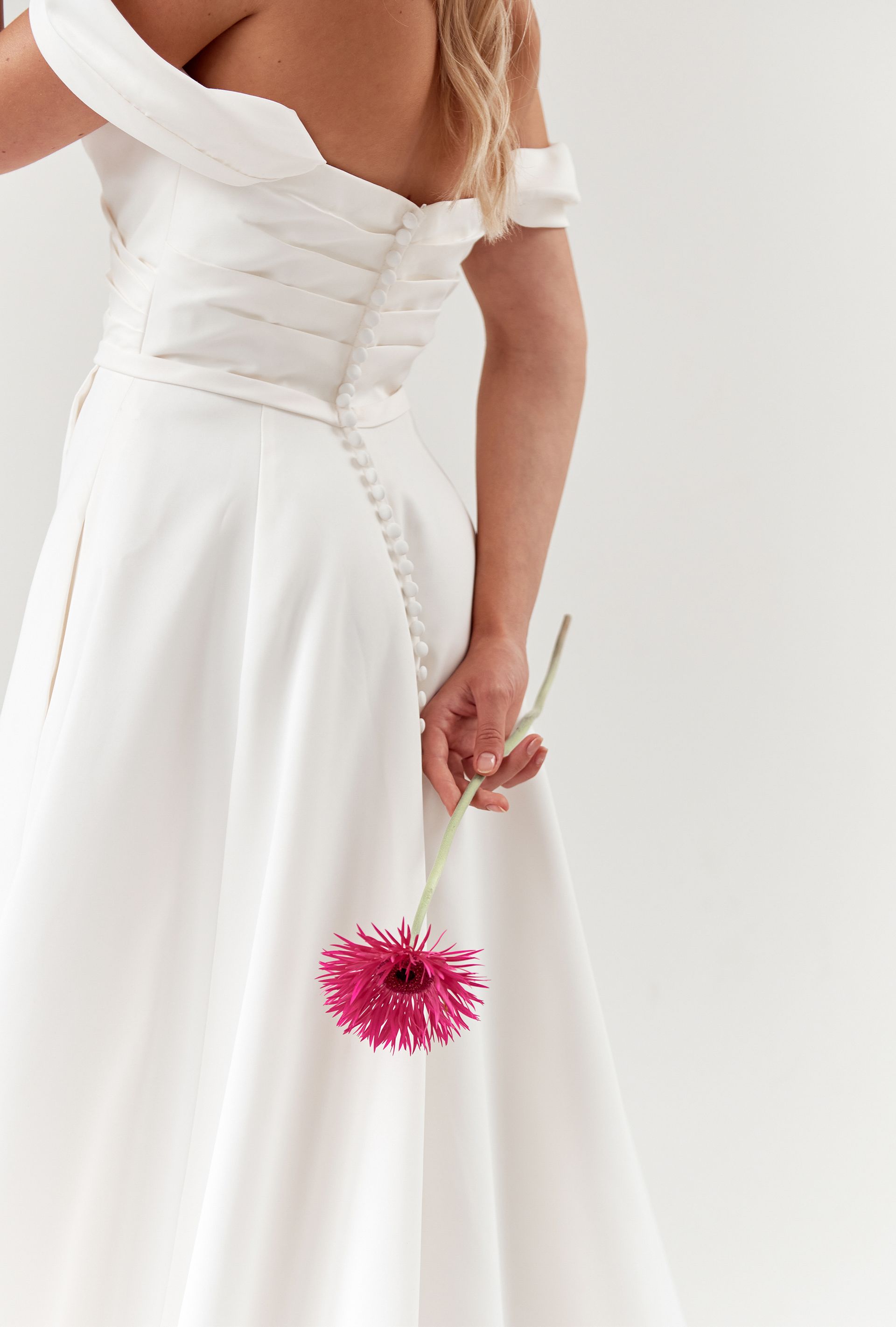 Woman in white off-the-shoulder dress, holding a pink flower behind her back, against a white background.