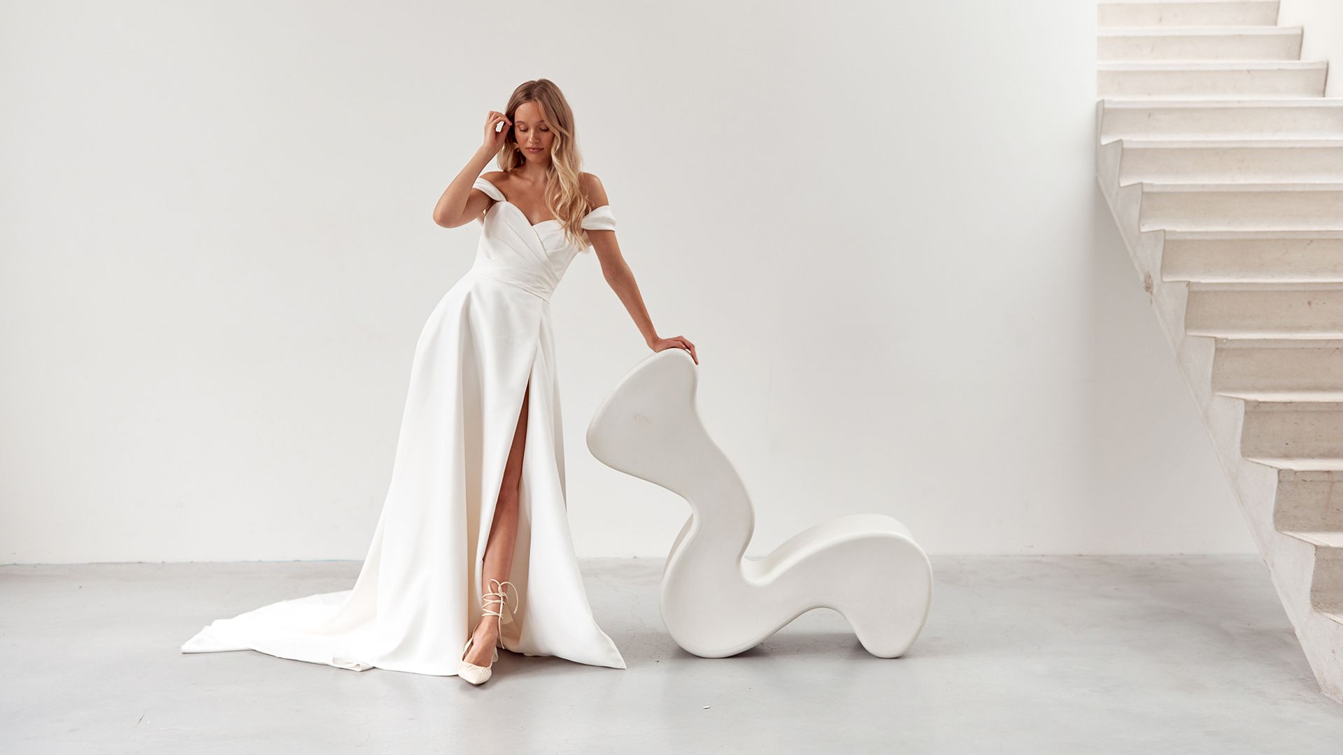 Woman in a white off-the-shoulder dress with high slit, leaning on a modern white chair near a staircase.