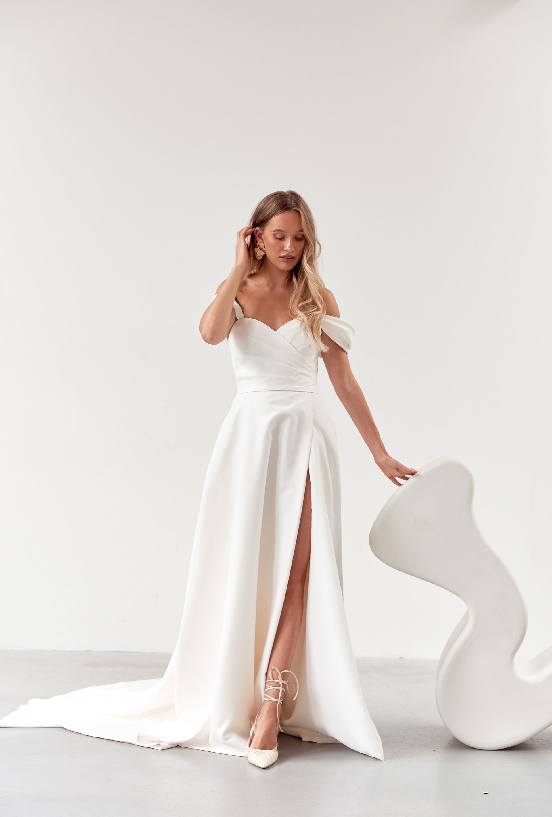 Woman in white wedding dress with high slit, standing indoors near a white sculptural element.