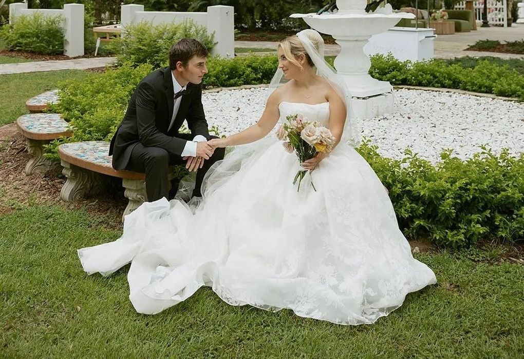 Bride and groom sitting by a fountain in a garden, holding hands, bride in white gown, groom in tuxedo.