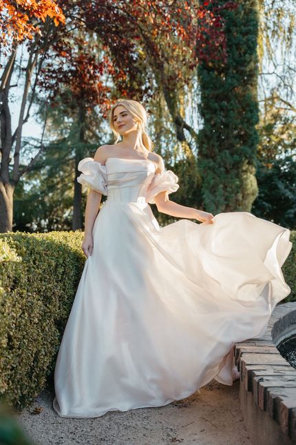 Woman in white off-shoulder gown, posing outdoors near greenery, with a flowing skirt.