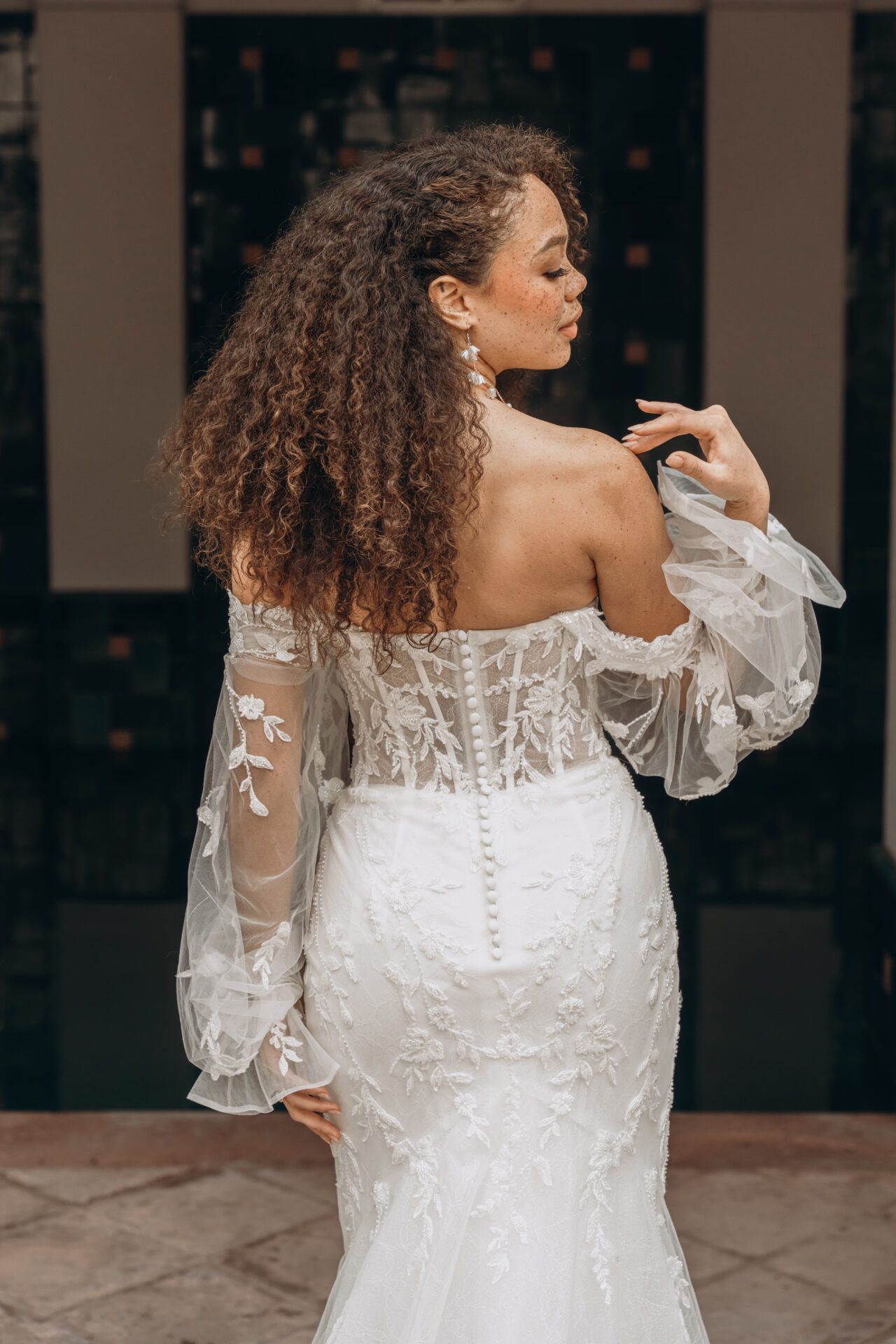 Woman in white off-shoulder wedding dress, curly hair, back to camera, hand on neck. Outdoors.