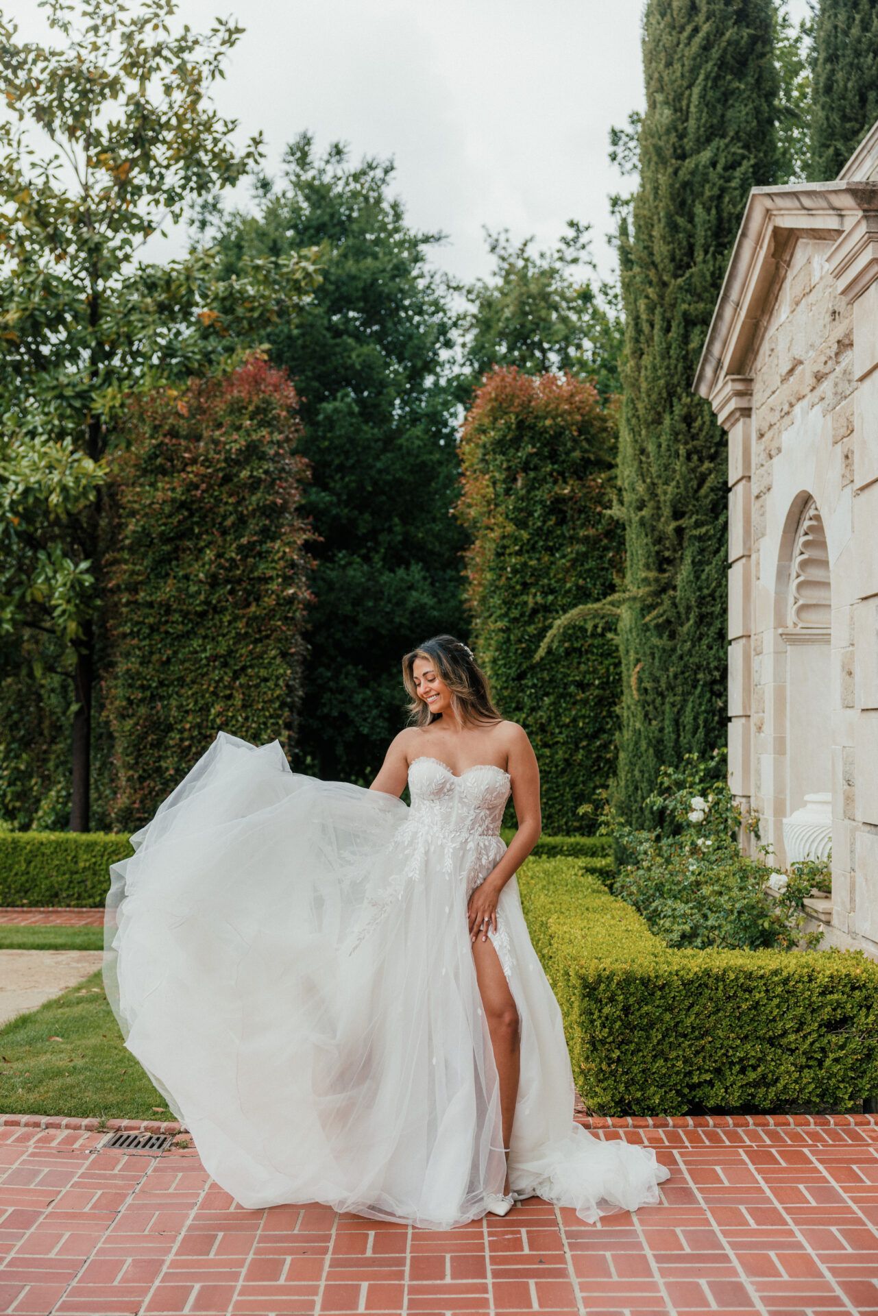 Woman in a flowing white gown with a high slit, posing outdoors near a building and greenery.