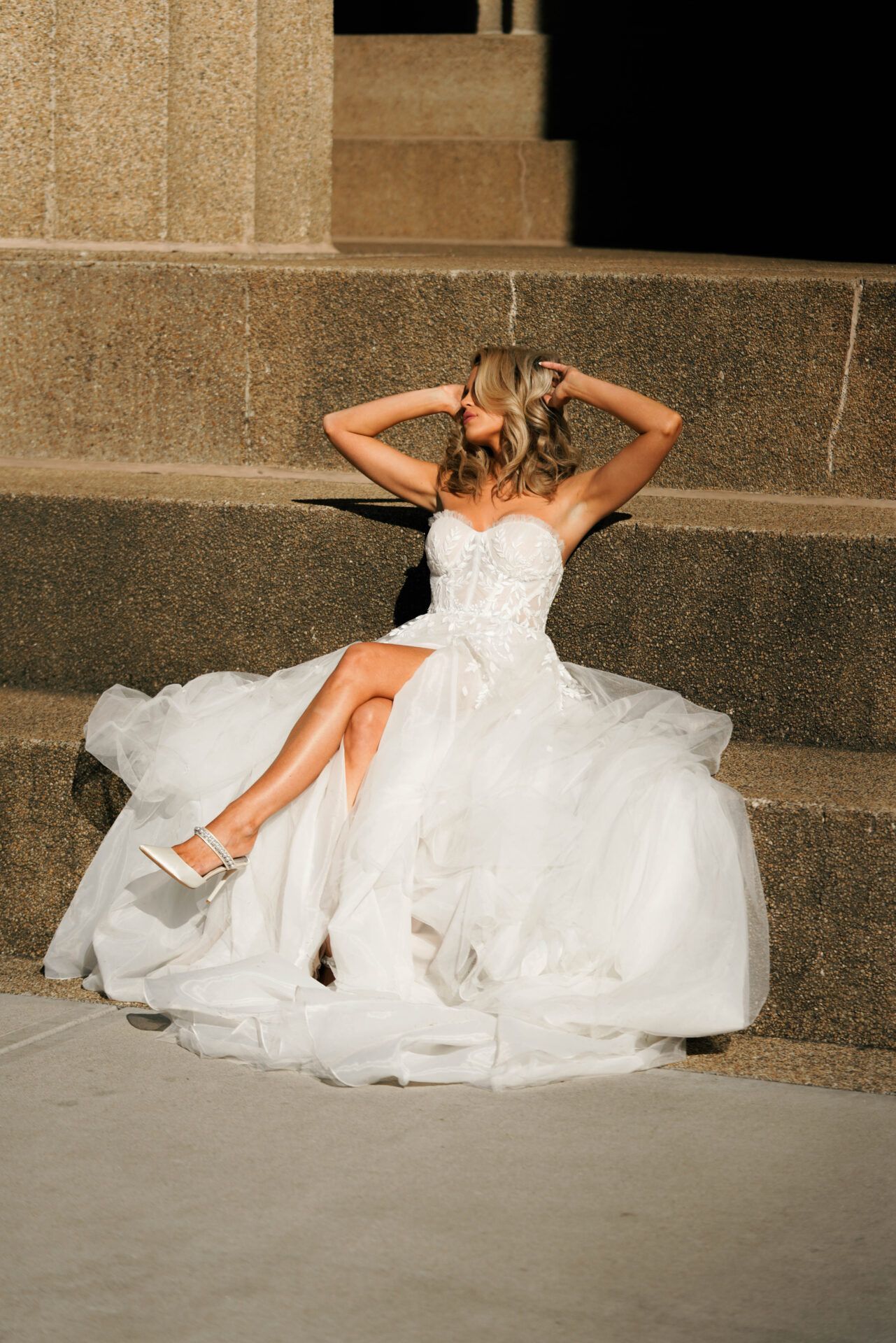 Woman in white wedding dress sits on outdoor steps, arms above head, leg crossed, sunny.