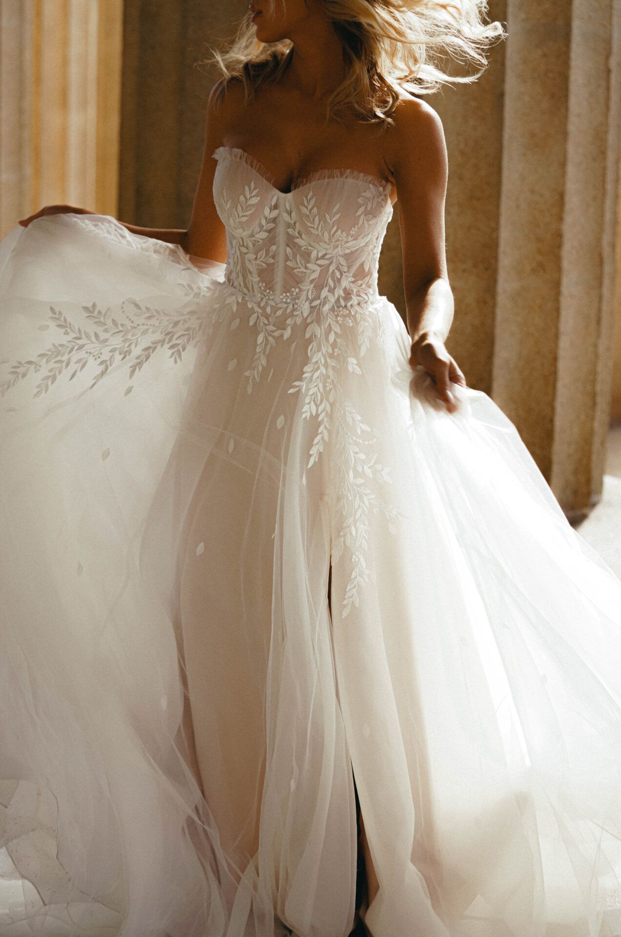 Woman in white strapless wedding gown with flowing skirt, standing in a building with columns, light streaming in.