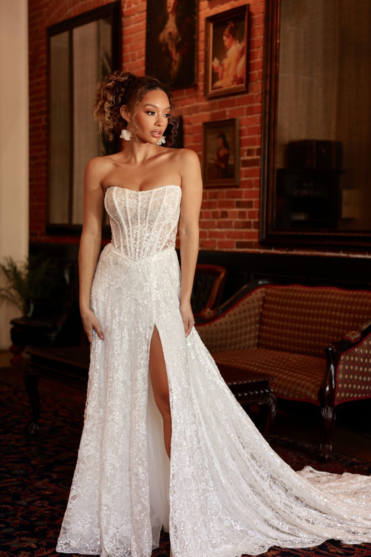 Woman in strapless white gown with high slit poses indoors; brick wall and art in background.