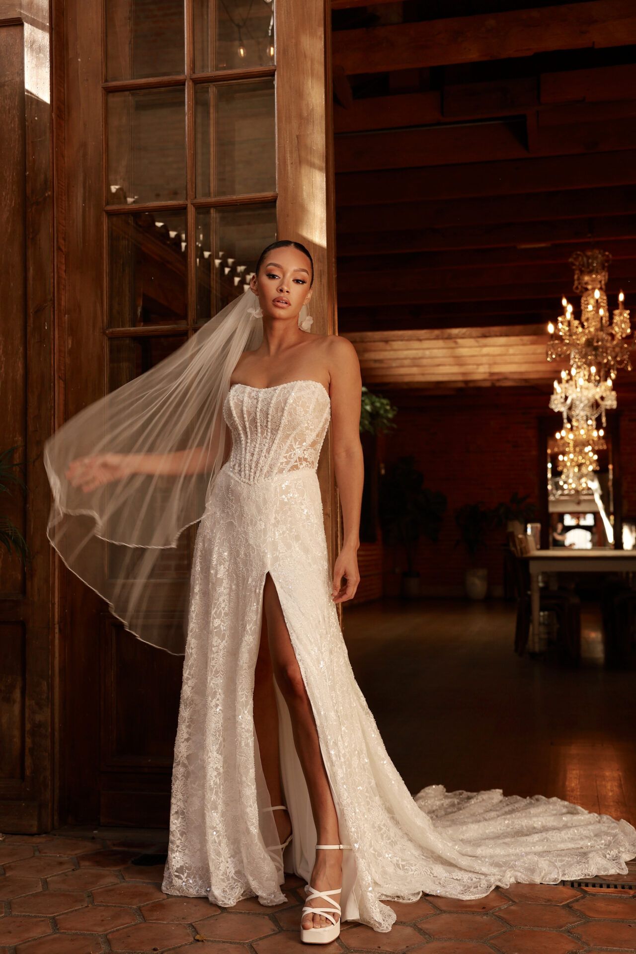 Woman in a sparkly white wedding dress with a high slit, veil, and strappy heels, standing in a wooden doorway.