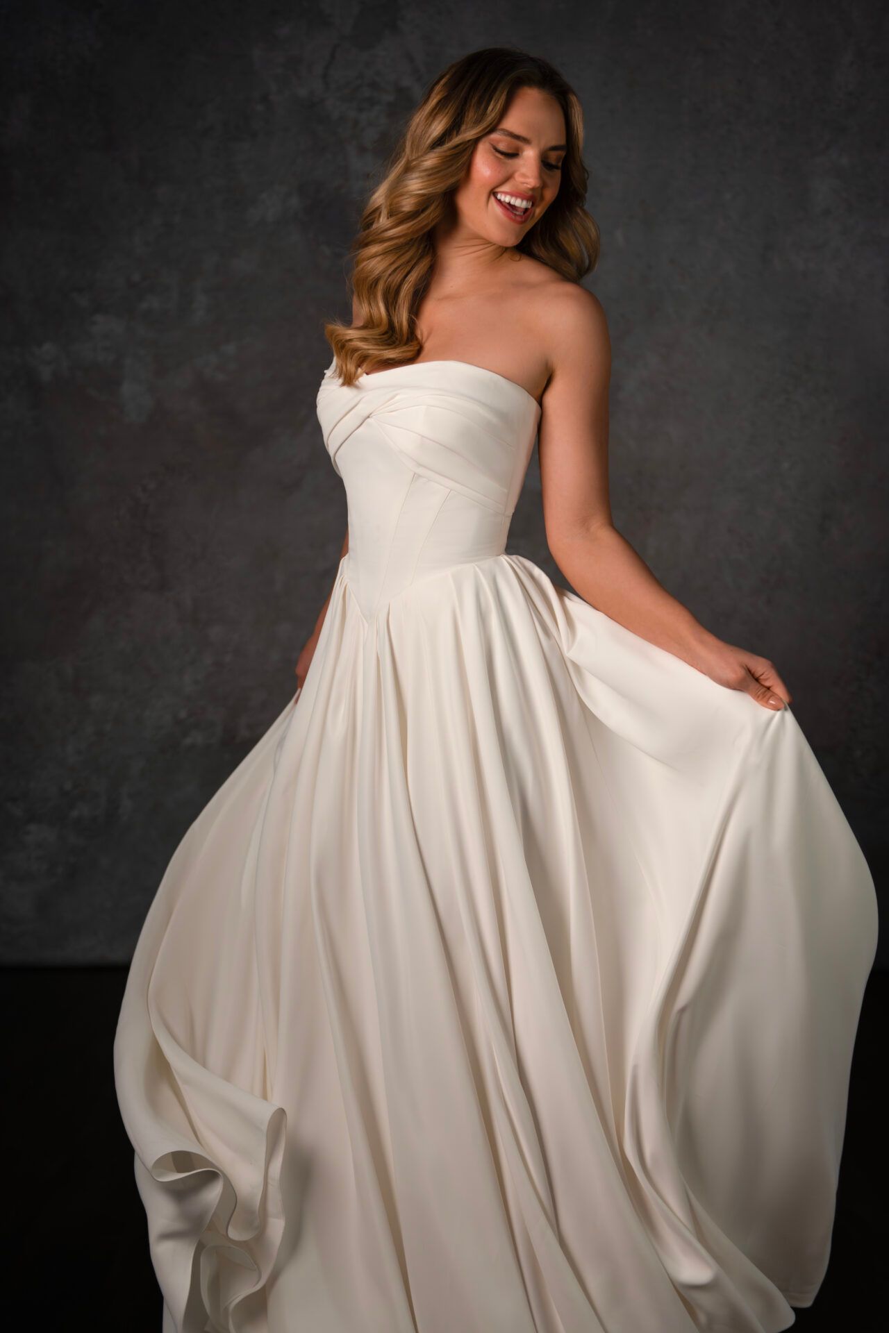 Woman in a white strapless gown smiles, holding up the skirt; against a dark gray backdrop.