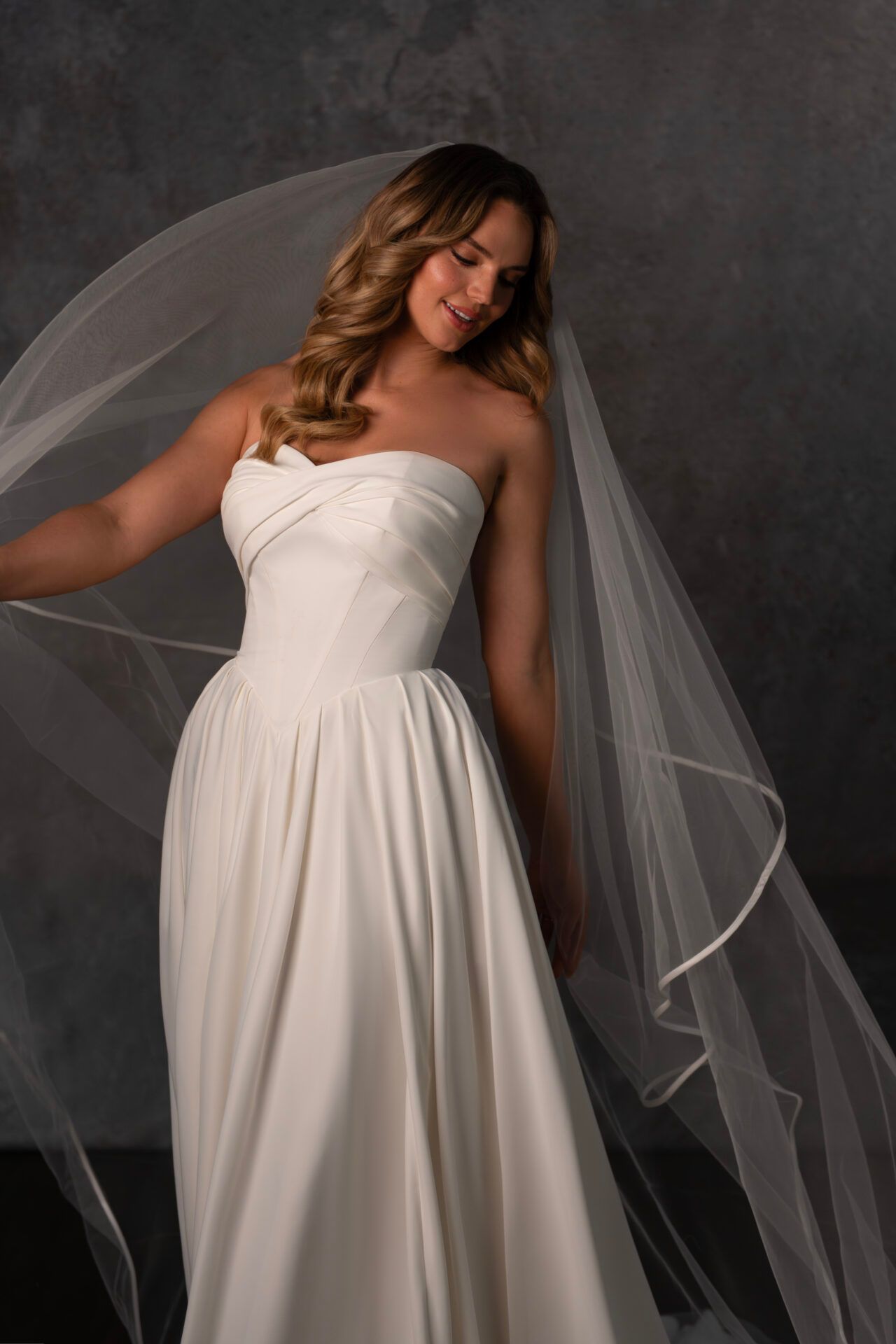 Woman in a strapless white wedding dress with a long veil, smiling and looking down.