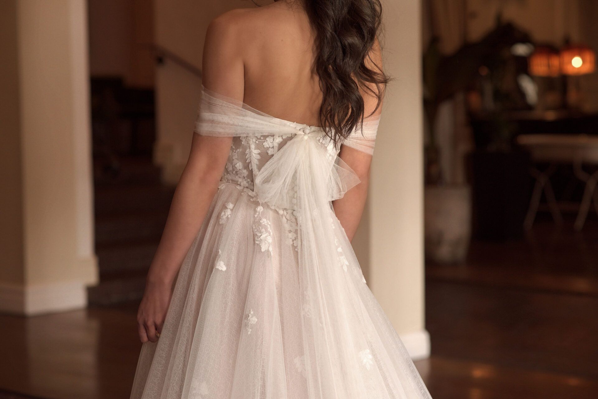 Woman in a white wedding dress, back view, standing indoors, tulle fabric with off-shoulder sleeves.