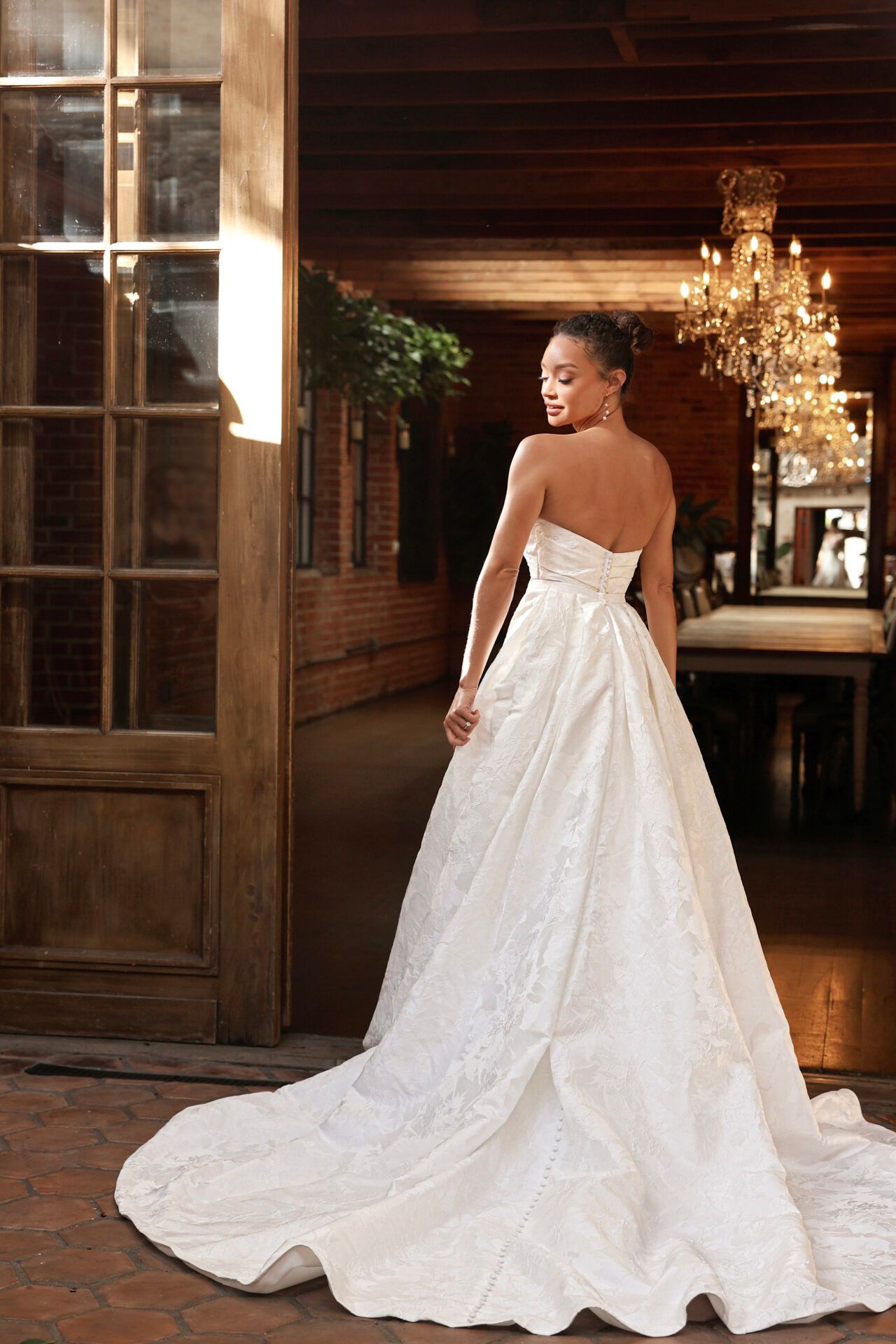 Woman in a white wedding dress stands by a wooden door, looking back. Indoor setting, chandelier.