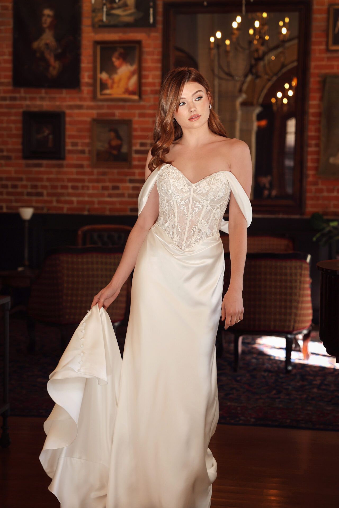 Woman in white off-the-shoulder gown holding fabric, standing in room with brick wall and paintings.