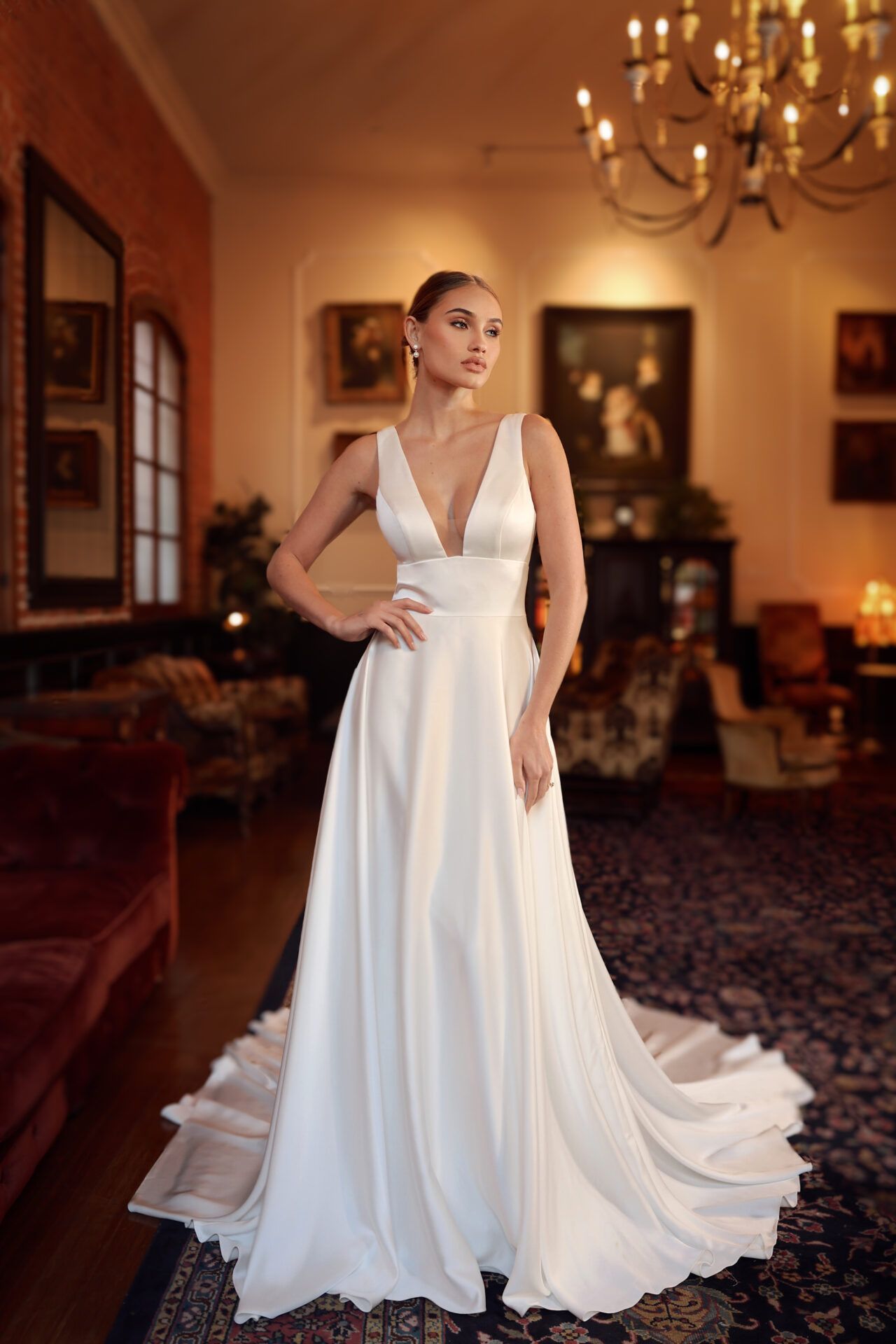 Woman in a white wedding gown, deep V-neckline, standing in a room with antique furniture and artwork.