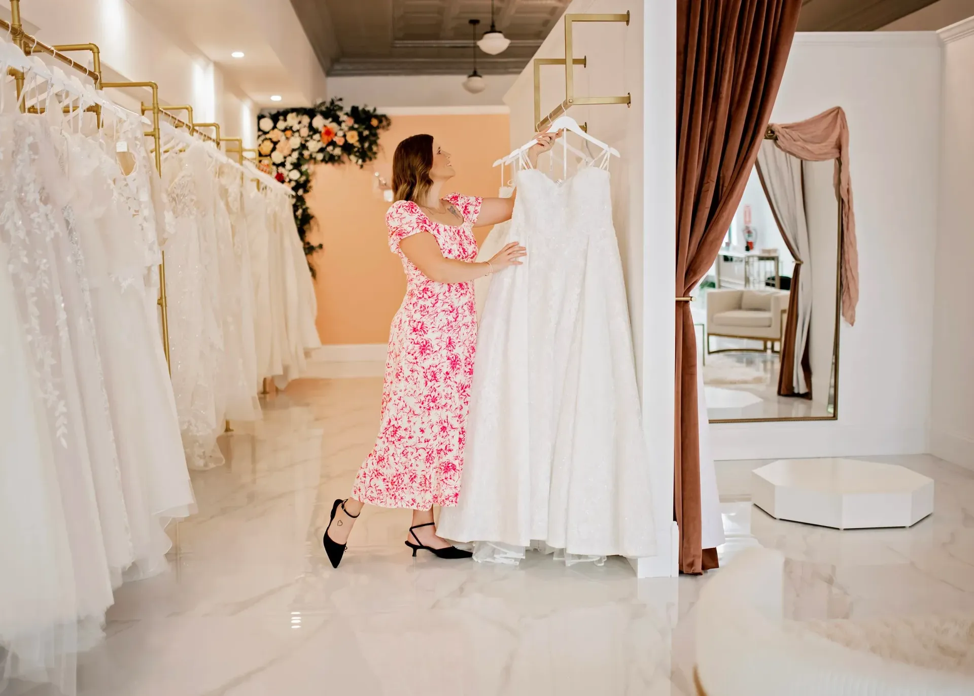 Woman in floral dress examines a white wedding dress in a bridal boutique.