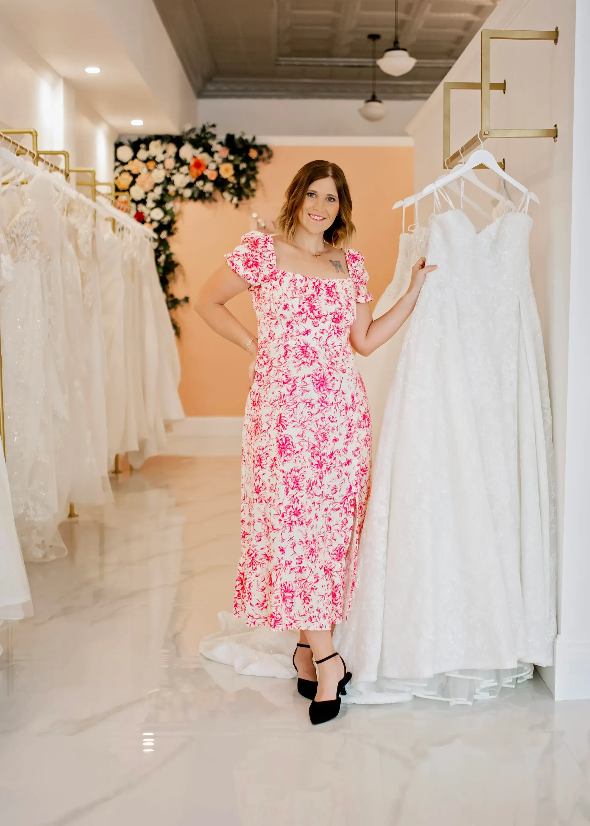 Woman in floral dress, smiles, holds up a wedding dress in a bridal shop.