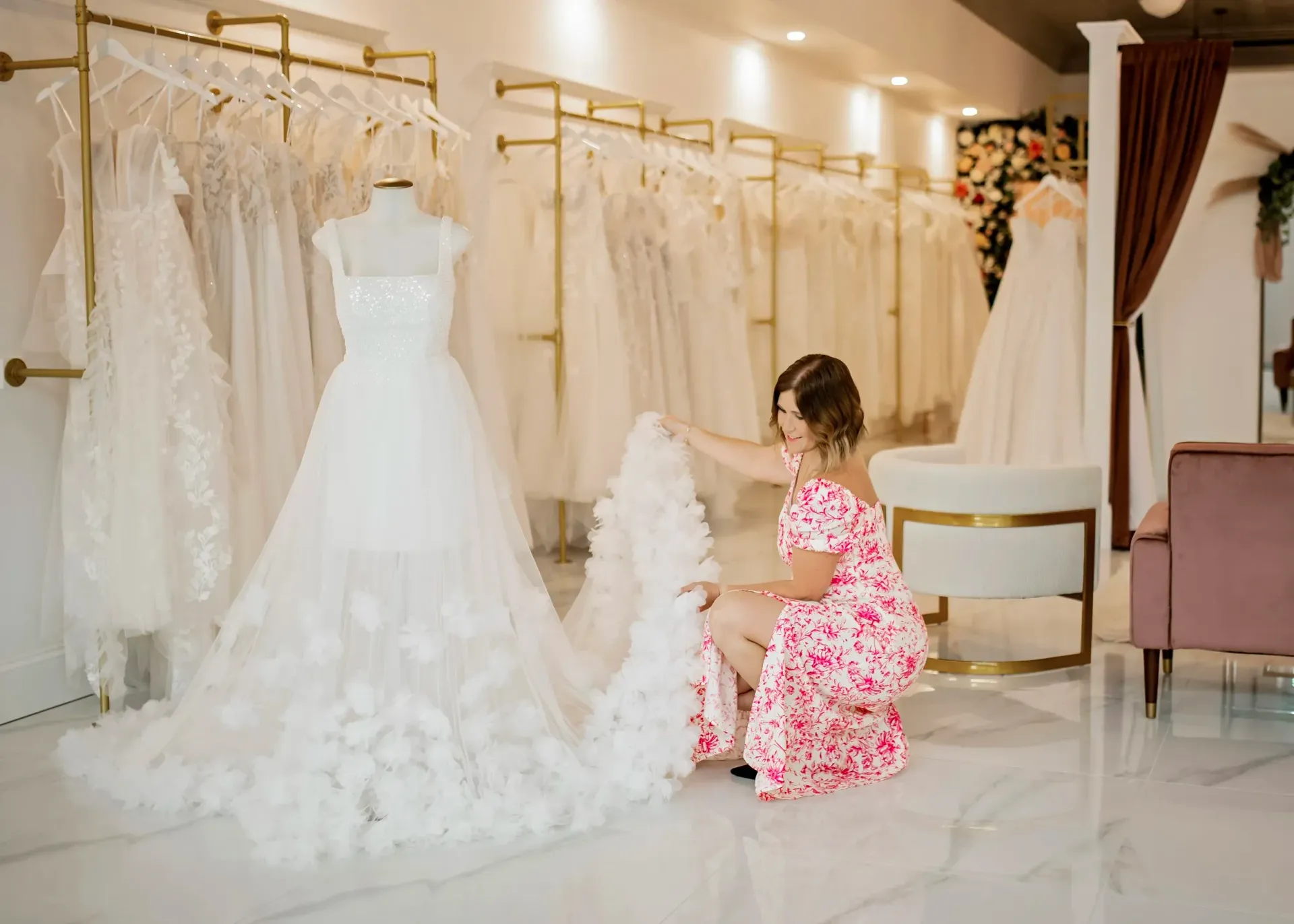 Woman adjusting a wedding dress on a mannequin in a bridal shop. Other dresses hang in the background.