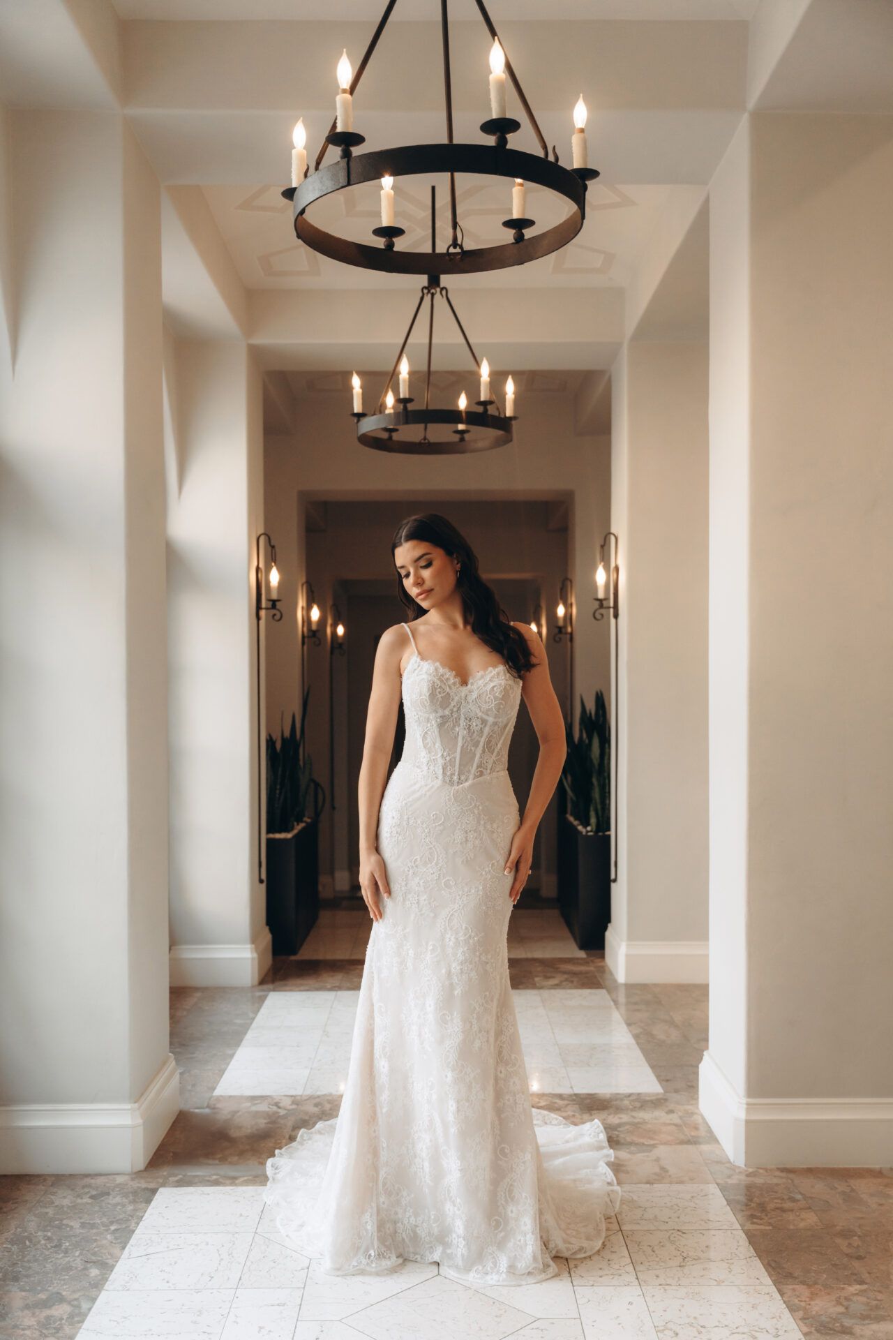 Bride in a fitted, beaded wedding dress poses in a hallway with chandeliers and sconces.