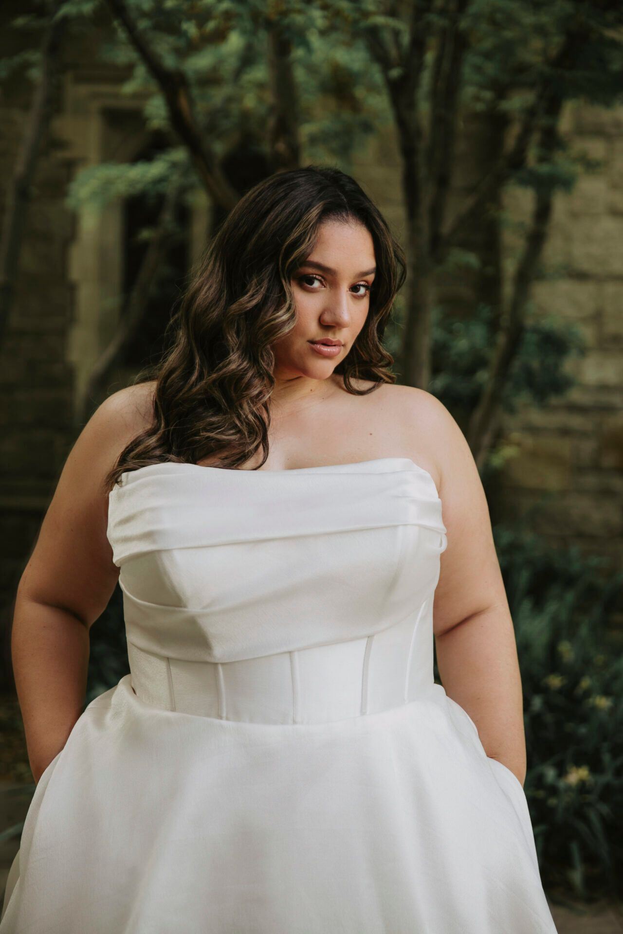Woman in a white strapless gown poses outdoors with hands in pockets; curly hair.
