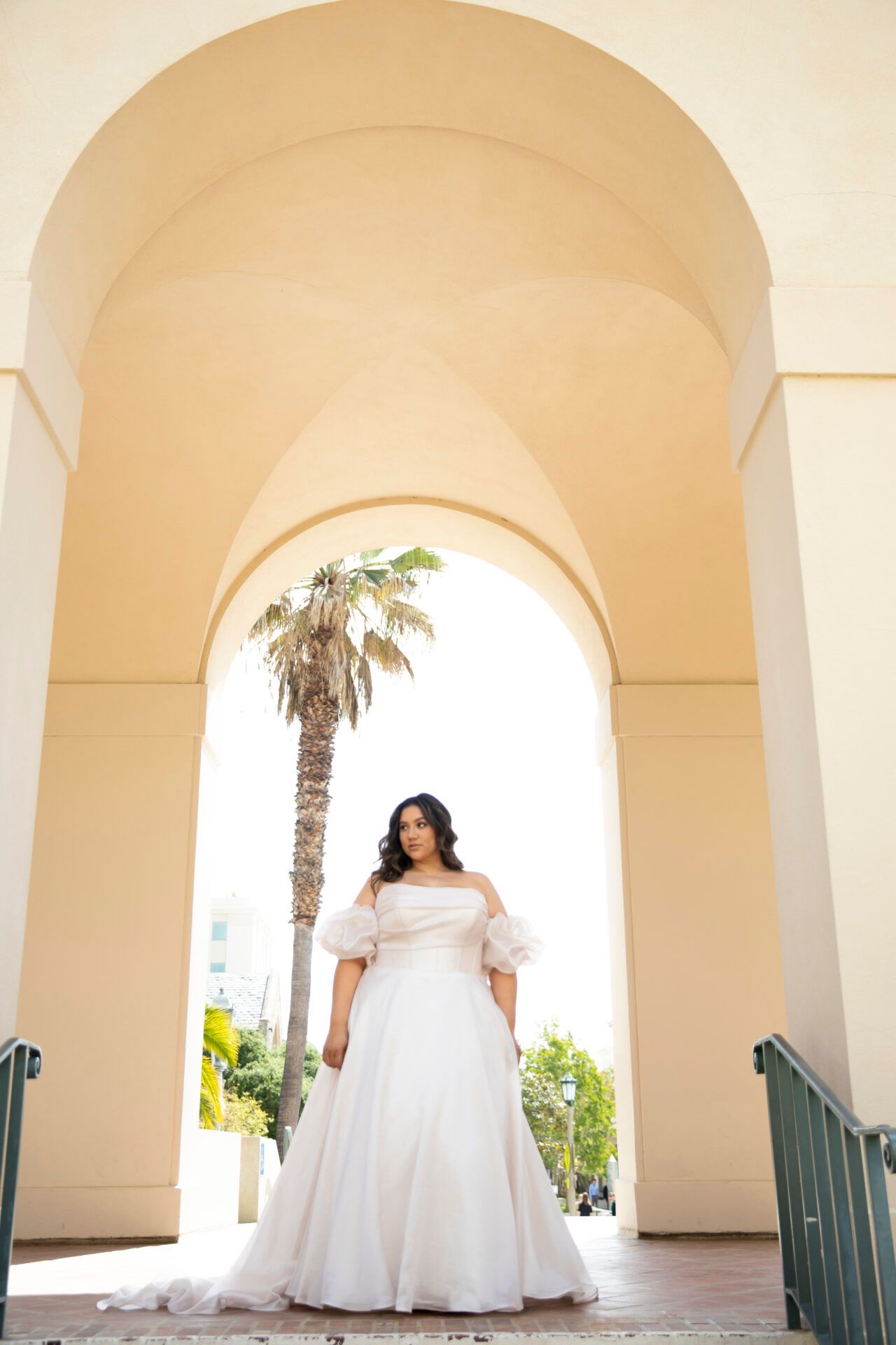 Woman in a pale pink off-shoulder gown stands under a beige arched doorway. A palm tree is visible in the background.