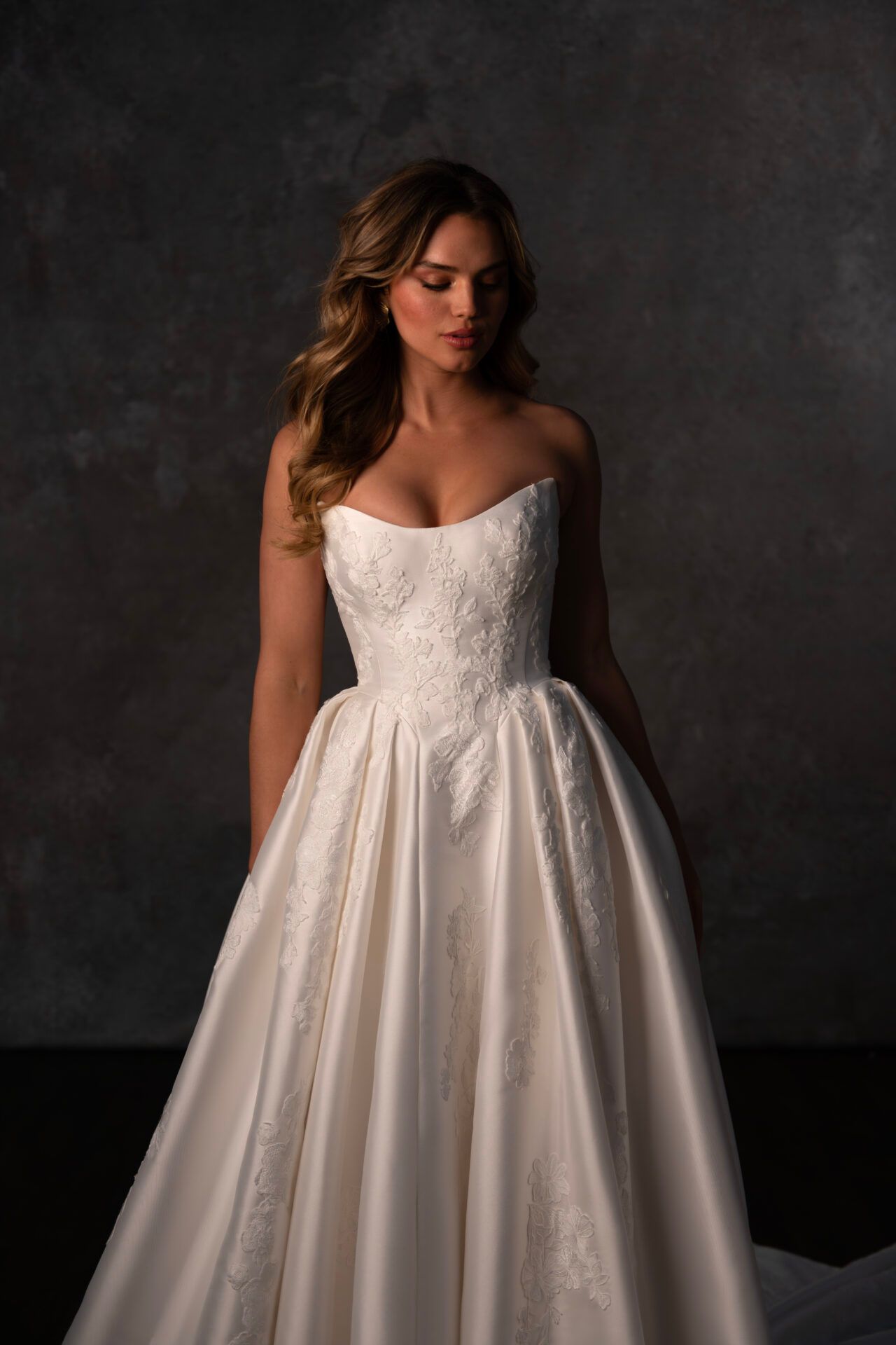 Woman in a strapless ivory wedding dress, looking down, in front of a dark backdrop.