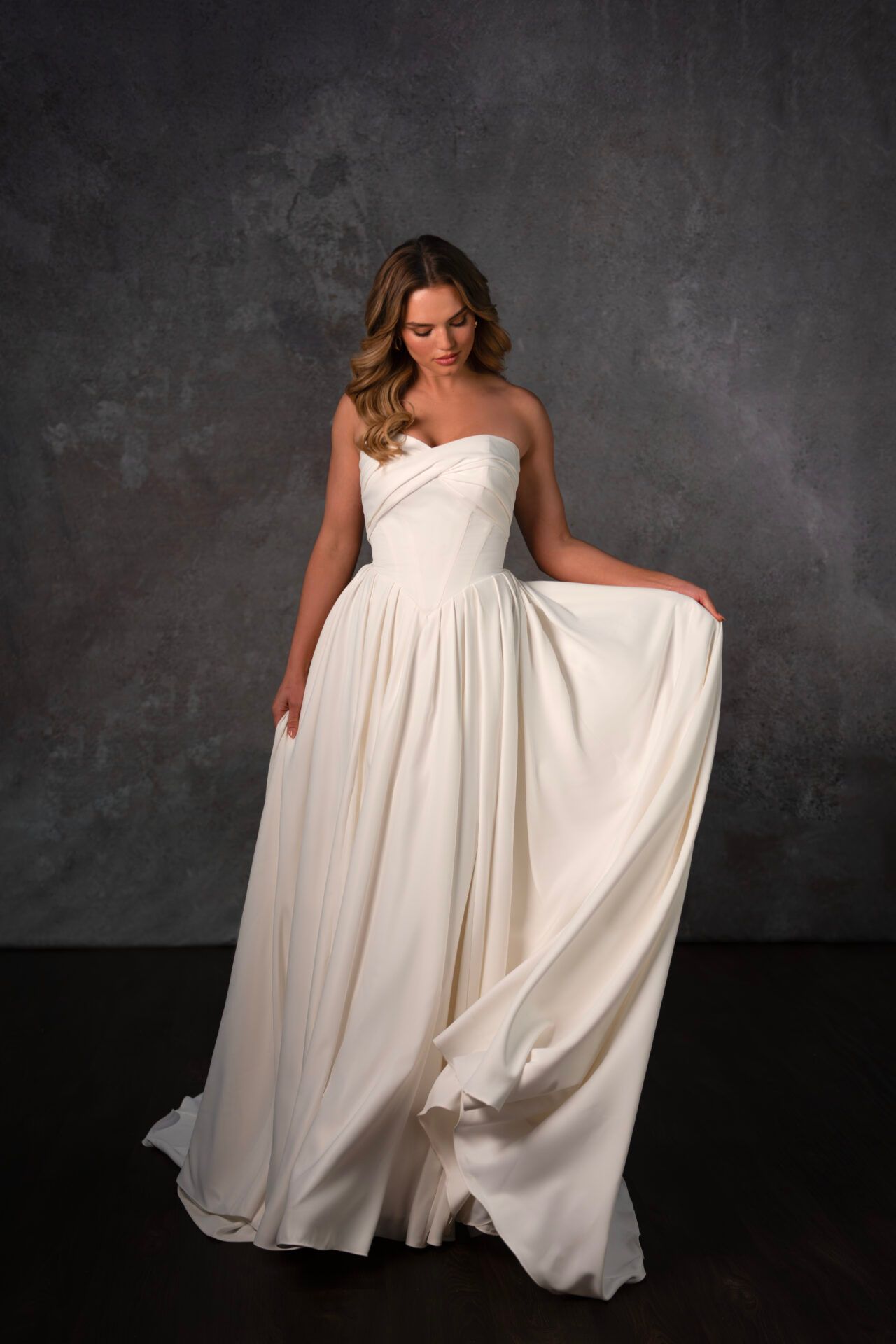 Woman in white strapless gown, holding up the skirt, stands against a gray background.