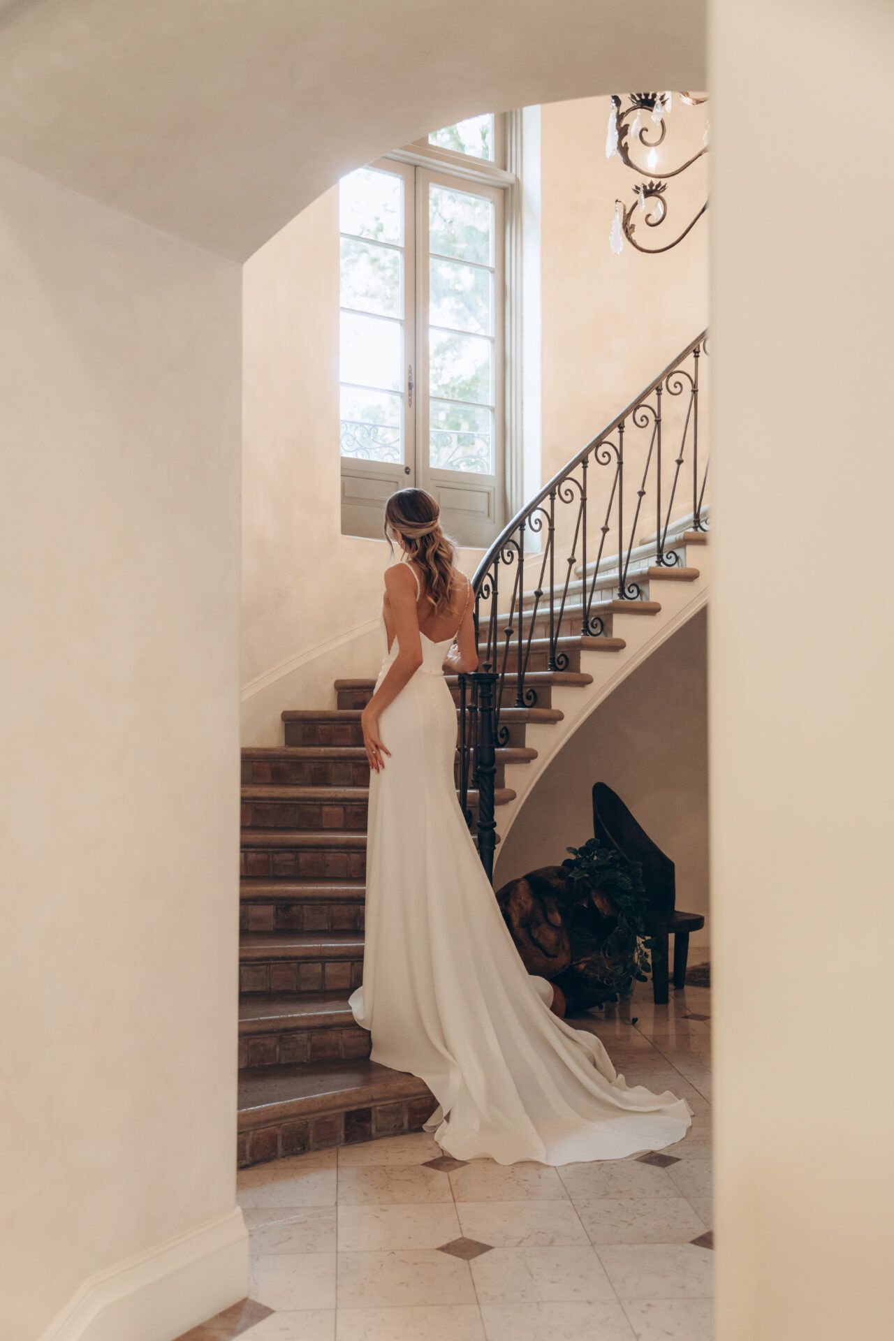 Bride in a white gown ascends a staircase, back to the camera, in a bright, ornate building.