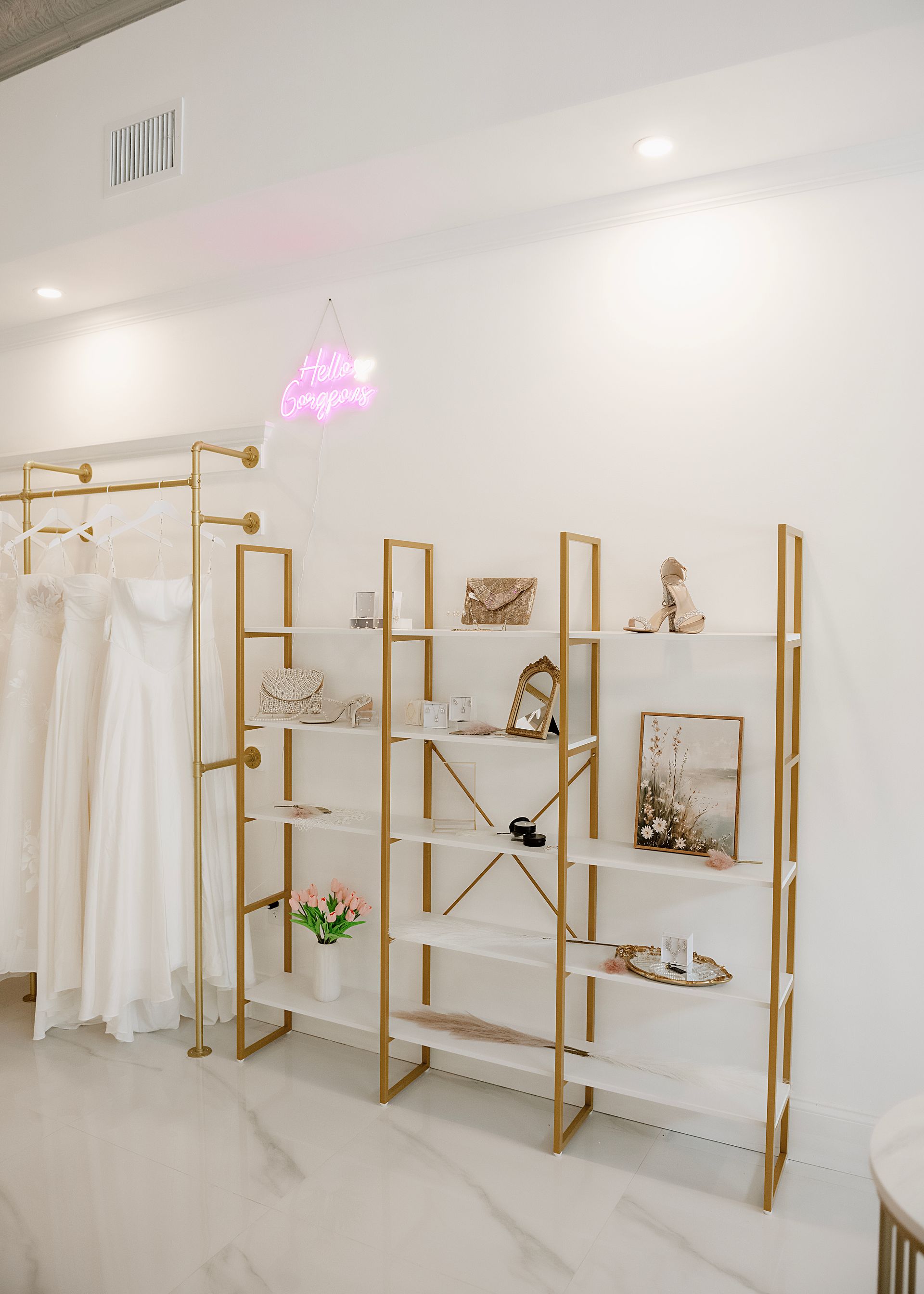 Bridal boutique interior with white dresses on rack, gold shelving displaying accessories, and a neon sign.