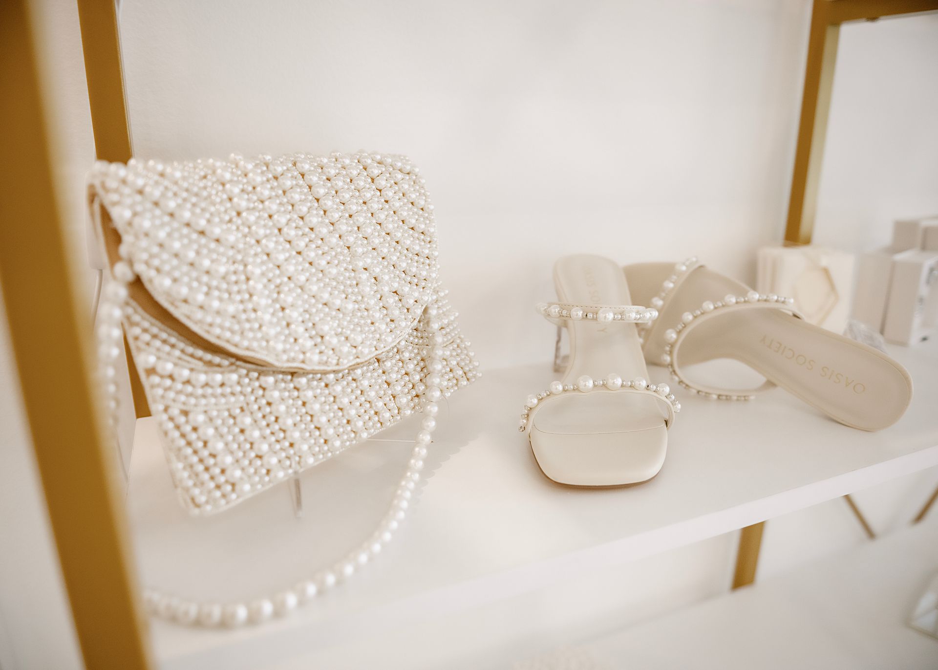 Pearl-covered handbag and matching sandals displayed on a white shelf. Beige tones.