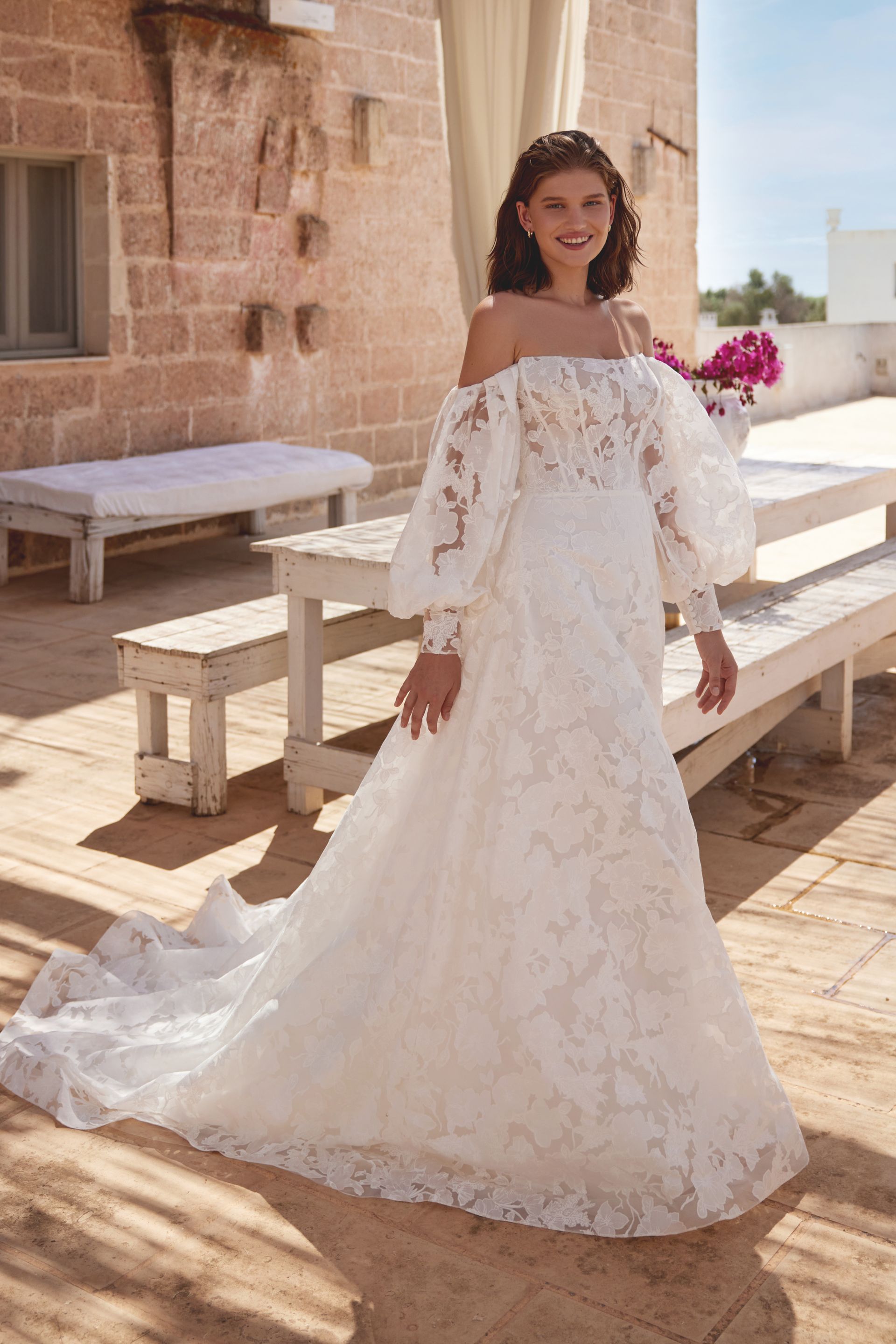 Woman in a white off-the-shoulder wedding gown standing outside near a brick building.
