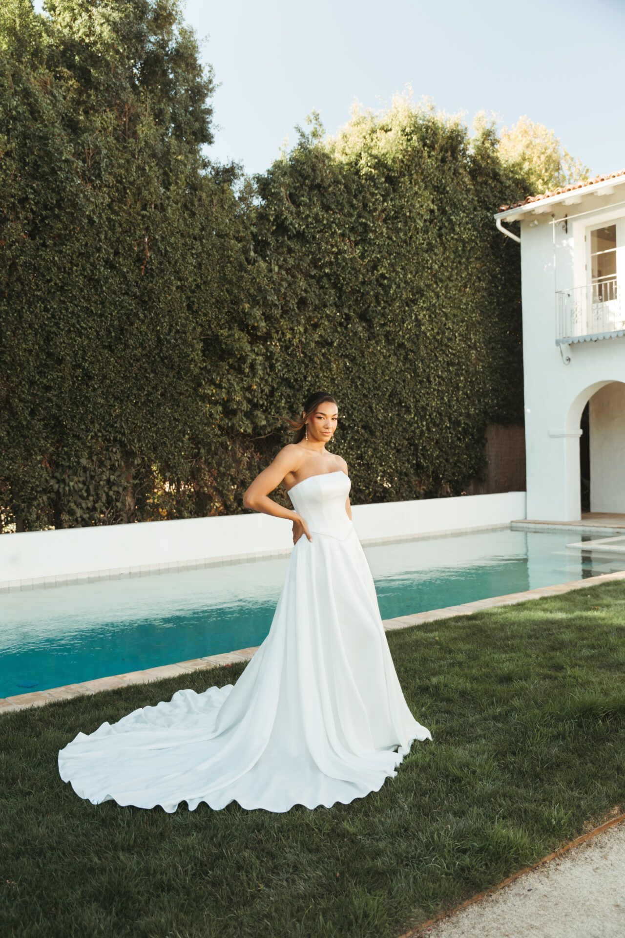 Woman in strapless wedding dress stands near pool, formal outdoor setting.
