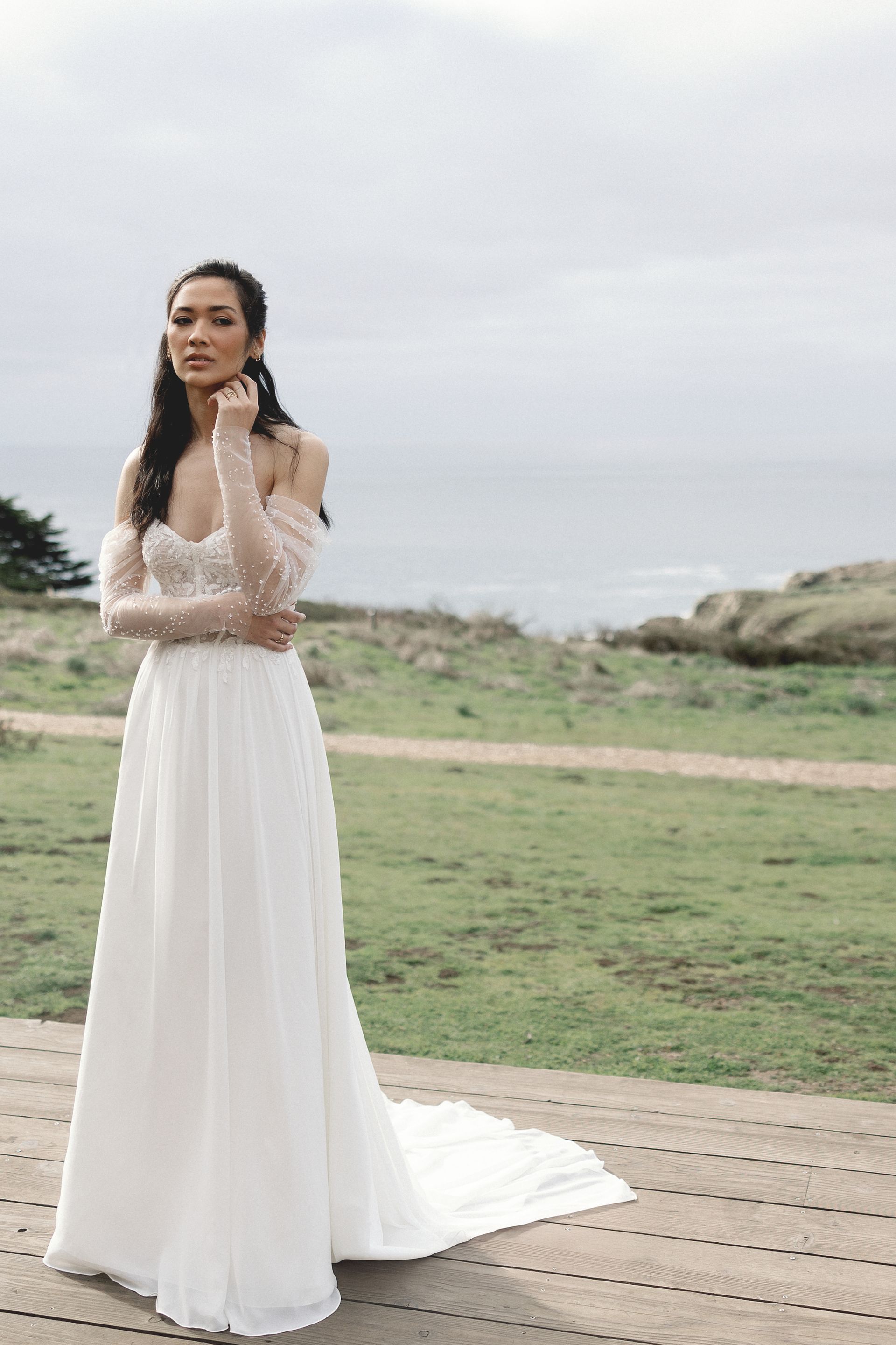 Woman in white off-shoulder wedding dress stands on a wooden deck overlooking the ocean.