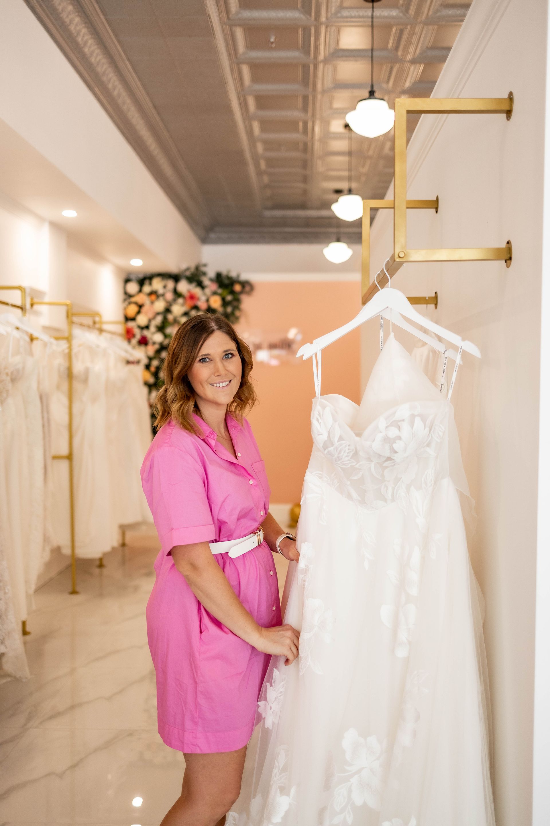 Woman in pink dress smiles beside a wedding dress in a bridal shop.