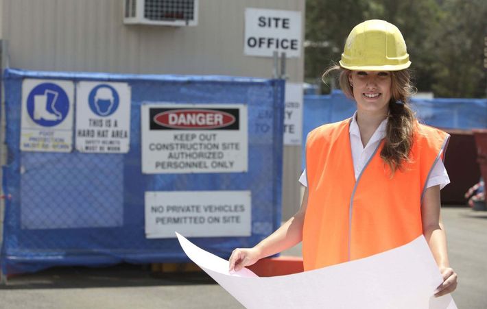 A woman wearing a hard hat is holding a blueprint in front of a danger sign