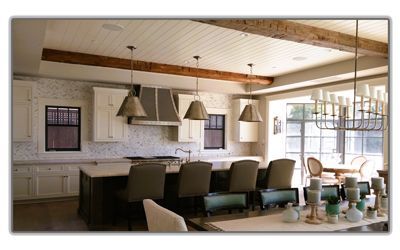 A spacious kitchen features a large dark island, light wood cabinets, three pendant lights, and wooden ceiling beams.