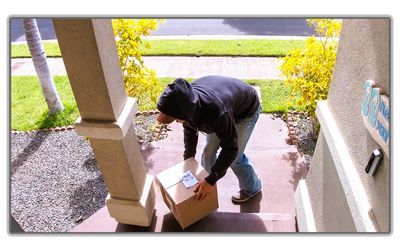 A person in a dark hooded sweatshirt picking up a package from a residential front porch.
