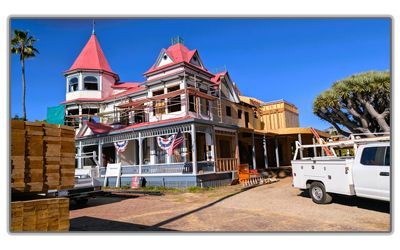 An ornate historic home with red roofs under construction, featuring wooden frames, a white truck, and a large tree nearby.