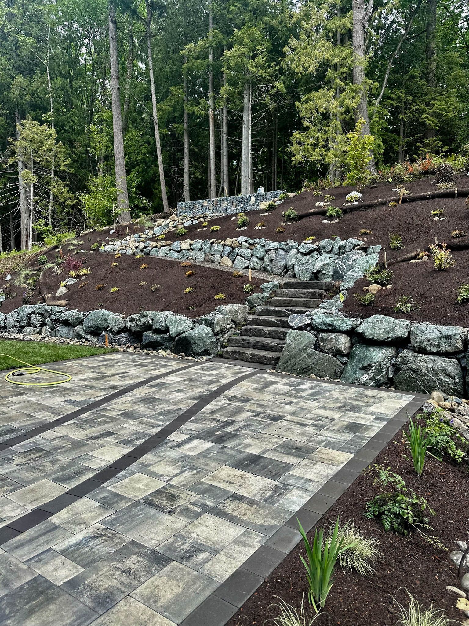 A patio with stairs leading up to a rock wall and trees in the background.