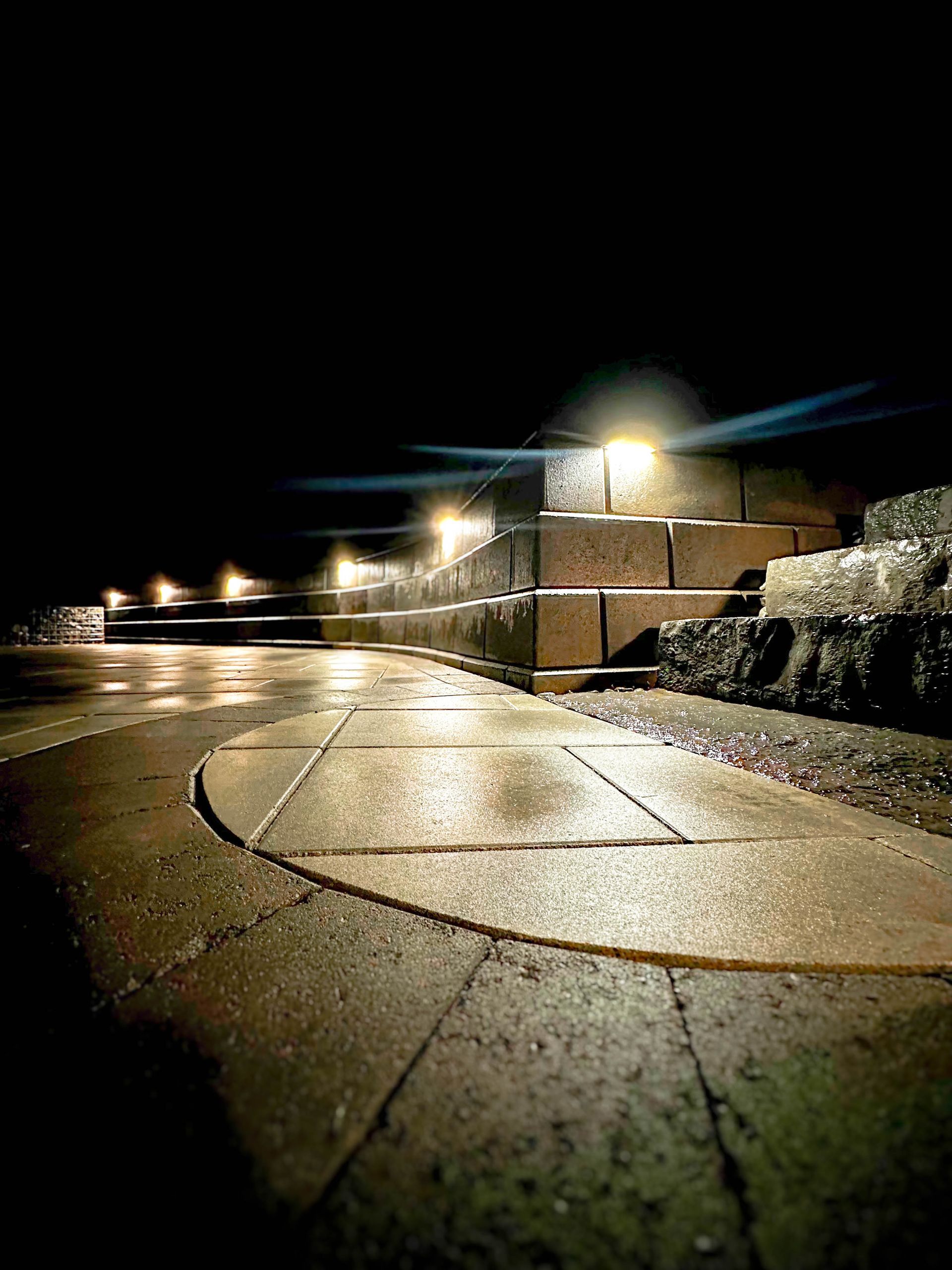 A brick wall is lit up at night along a sidewalk.
