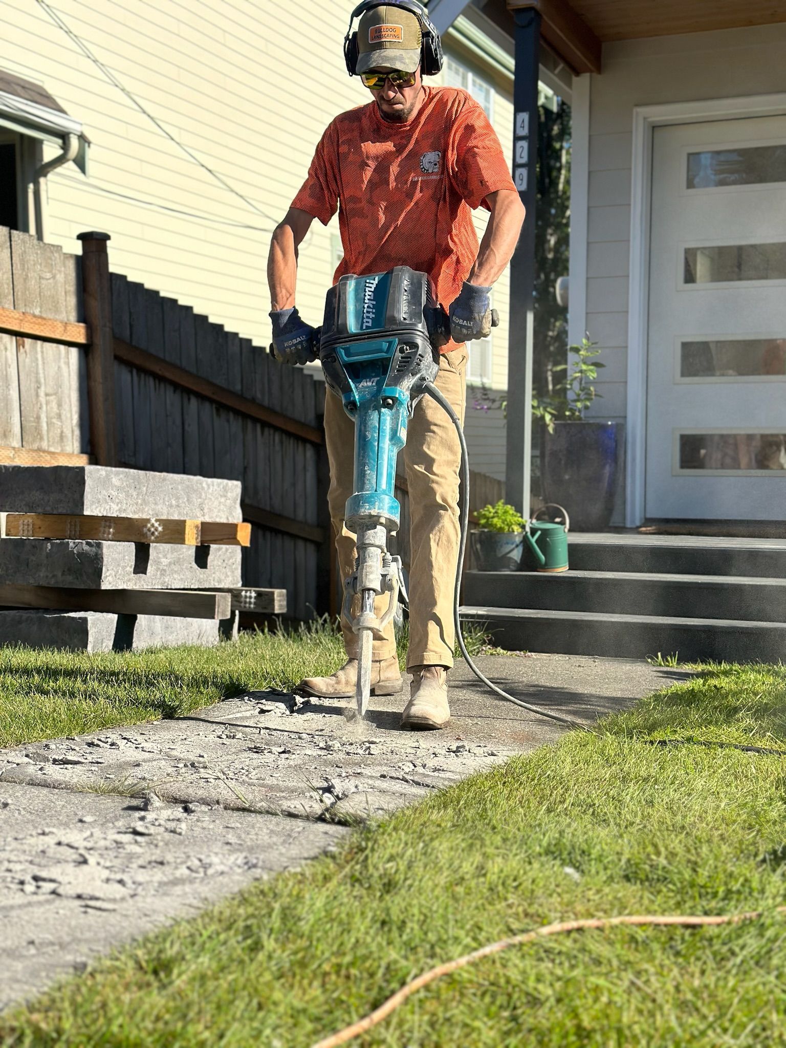 A man is using a concrete breaker on a sidewalk in front of a house.